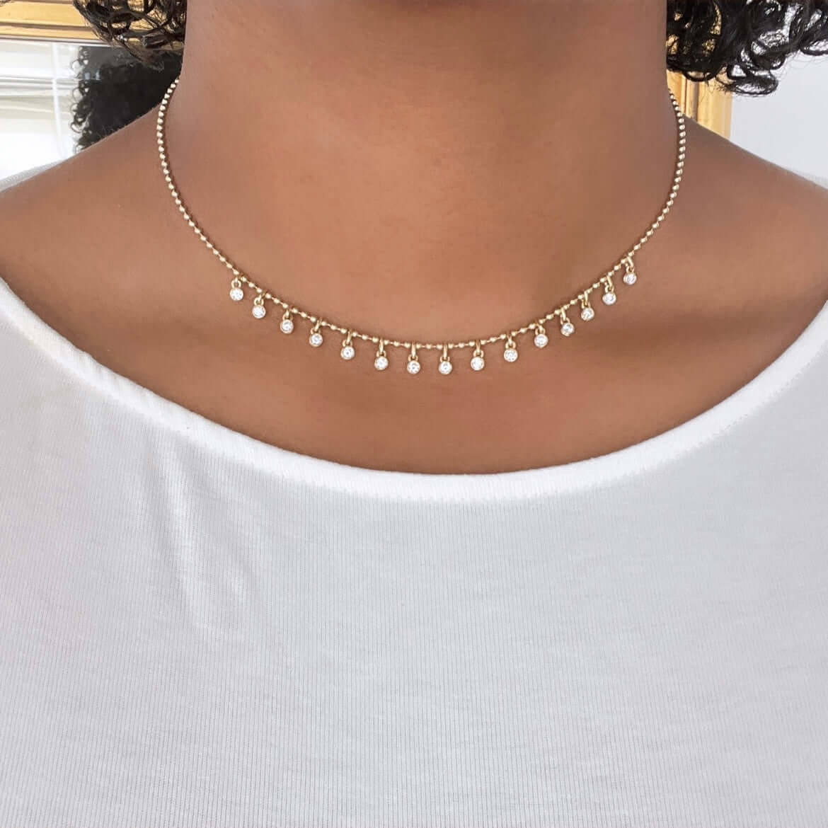 A close-up image shows a person wearing the Janet Heller Fine Jewelry Diamond Dallas Necklace, with small dangling white stones. The delicate piece adorns their neck above a white round-neck top, while the softly blurred background enhances its allure.