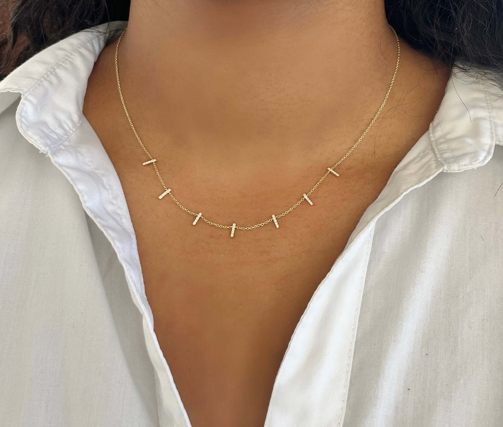 A person in a white open-collar shirt elegantly wears the Diamond Dangle Bar Necklace from Janet Heller Fine Jewelry, featuring small gold vertical bars on a neutral background.