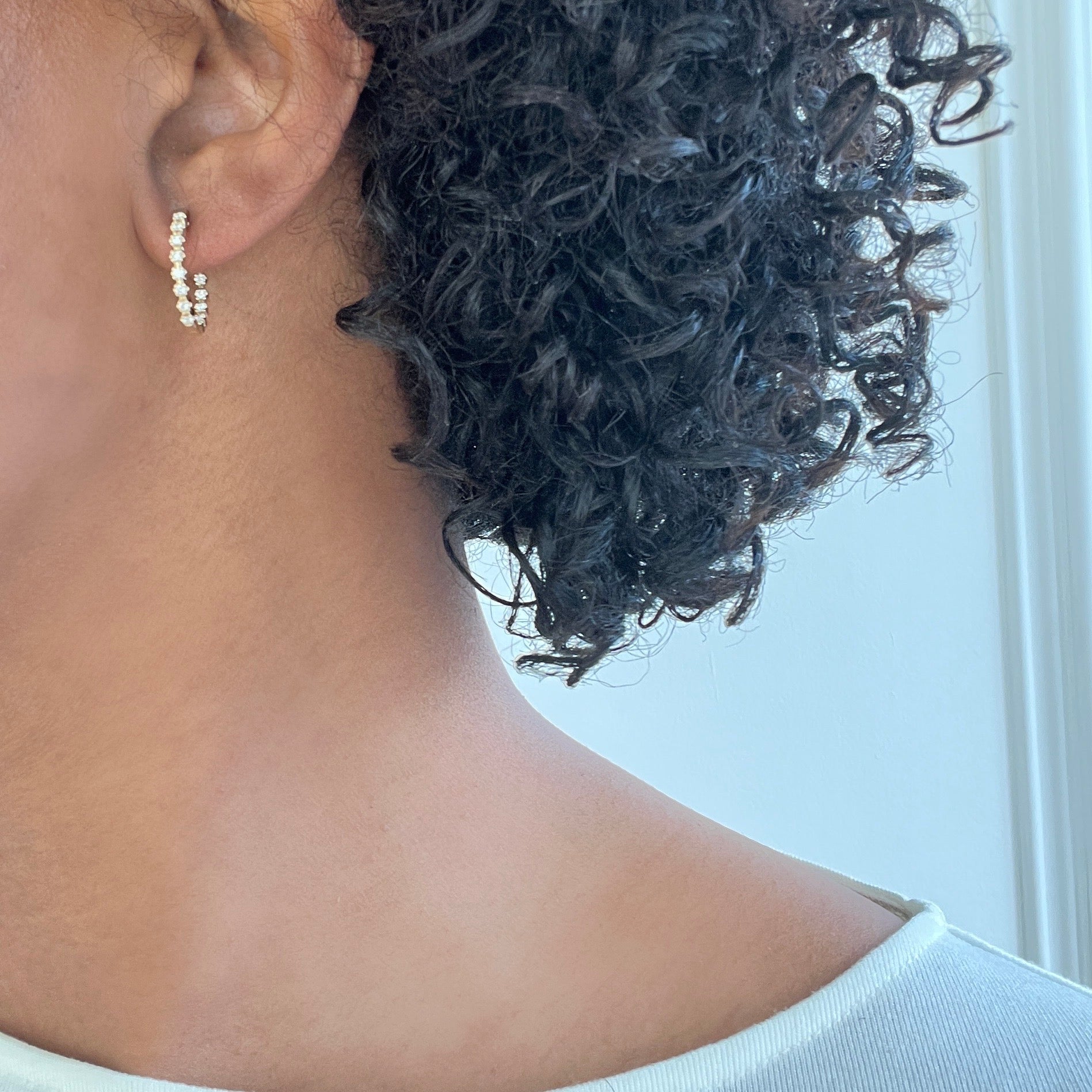 A close-up shows a person with curly hair wearing the elegant Diamond Leighton Hoops from Janet Heller Fine Jewelry, adorned with gemstones. The focus is on the ear and earring, partially veiled by hair against a softly lit background.