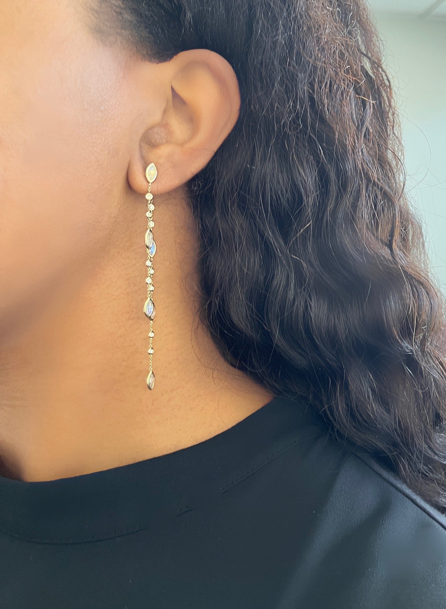 A close-up of someone with long, wavy dark hair in a black top showcases FoundryFineJewelrys Diamond & Moonstone Dangle Earrings. The image highlights the earrings elegant linked gemstones against a softly blurred background.