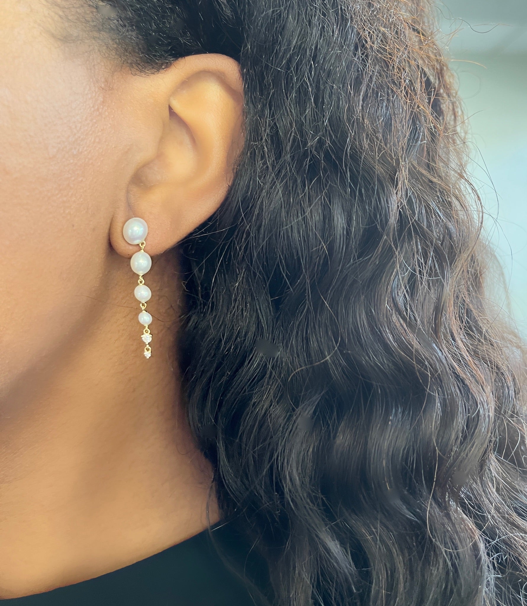 A persons ear, adorned with Janet Heller Fine Jewelrys exquisite Diamond & Pearl Drop Earrings, peeks through their long, wavy hair. The warm-toned skin contrasts with the softly blurred background, inviting inquiries about customizations for these timeless pieces.