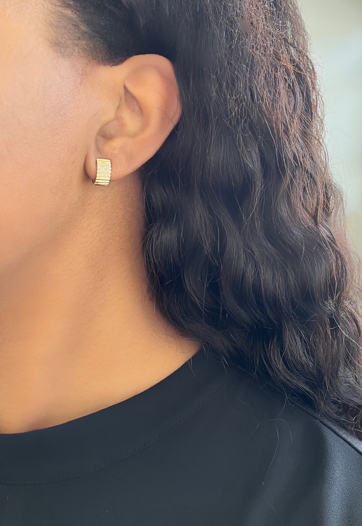 A person with long, wavy black hair wears a Diamond Ridge Huggie from Janet Heller Fine Jewelry. The small gold earrings textured surface adds exquisite detail. Dressed in a black top, only their side profile from the ear down is visible.