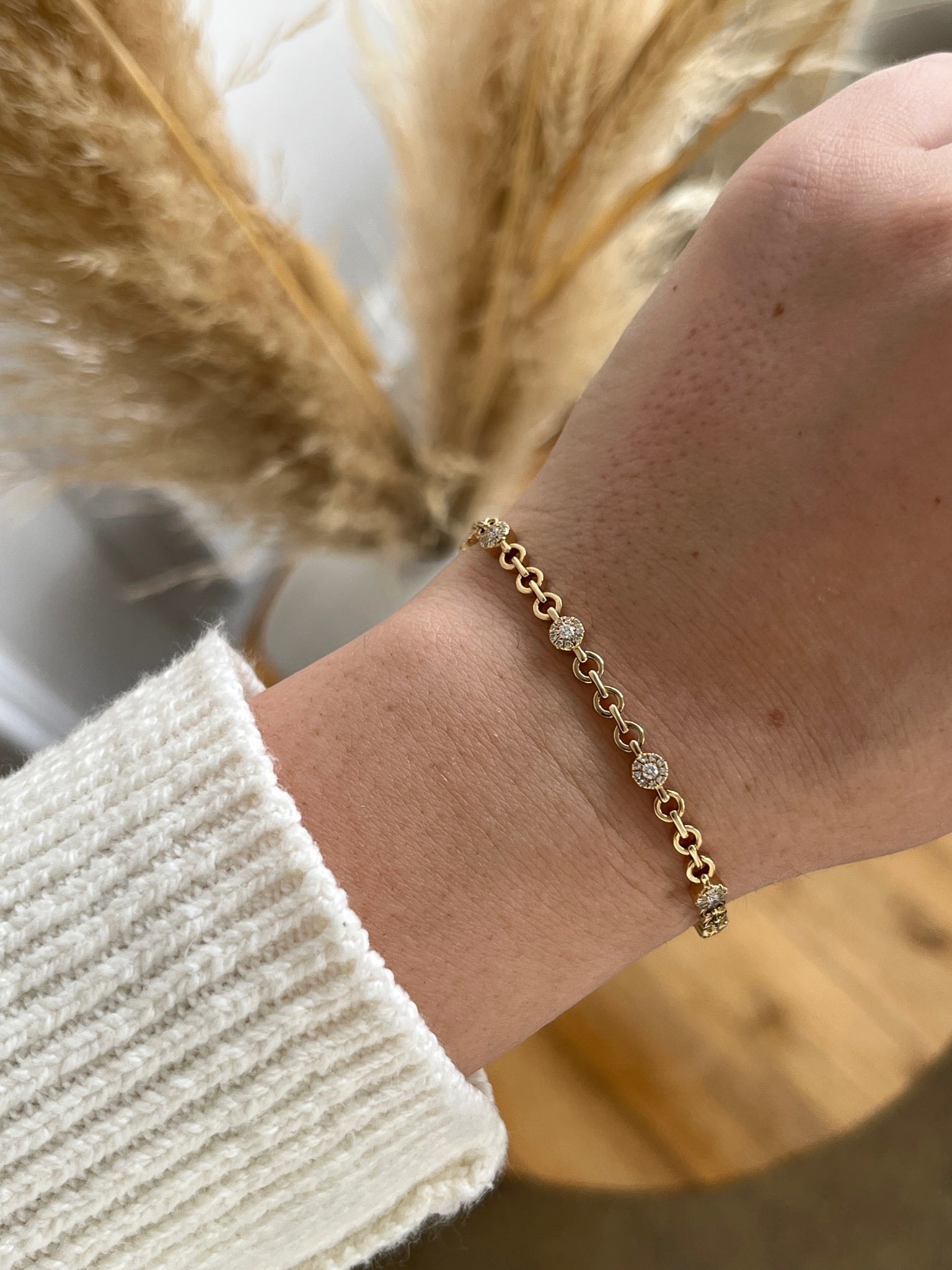 A hand adorned with the Diamond Round Link Bracelet rests gracefully against a white knitted sweater, framed softly by dried pampas grass stems in the background.