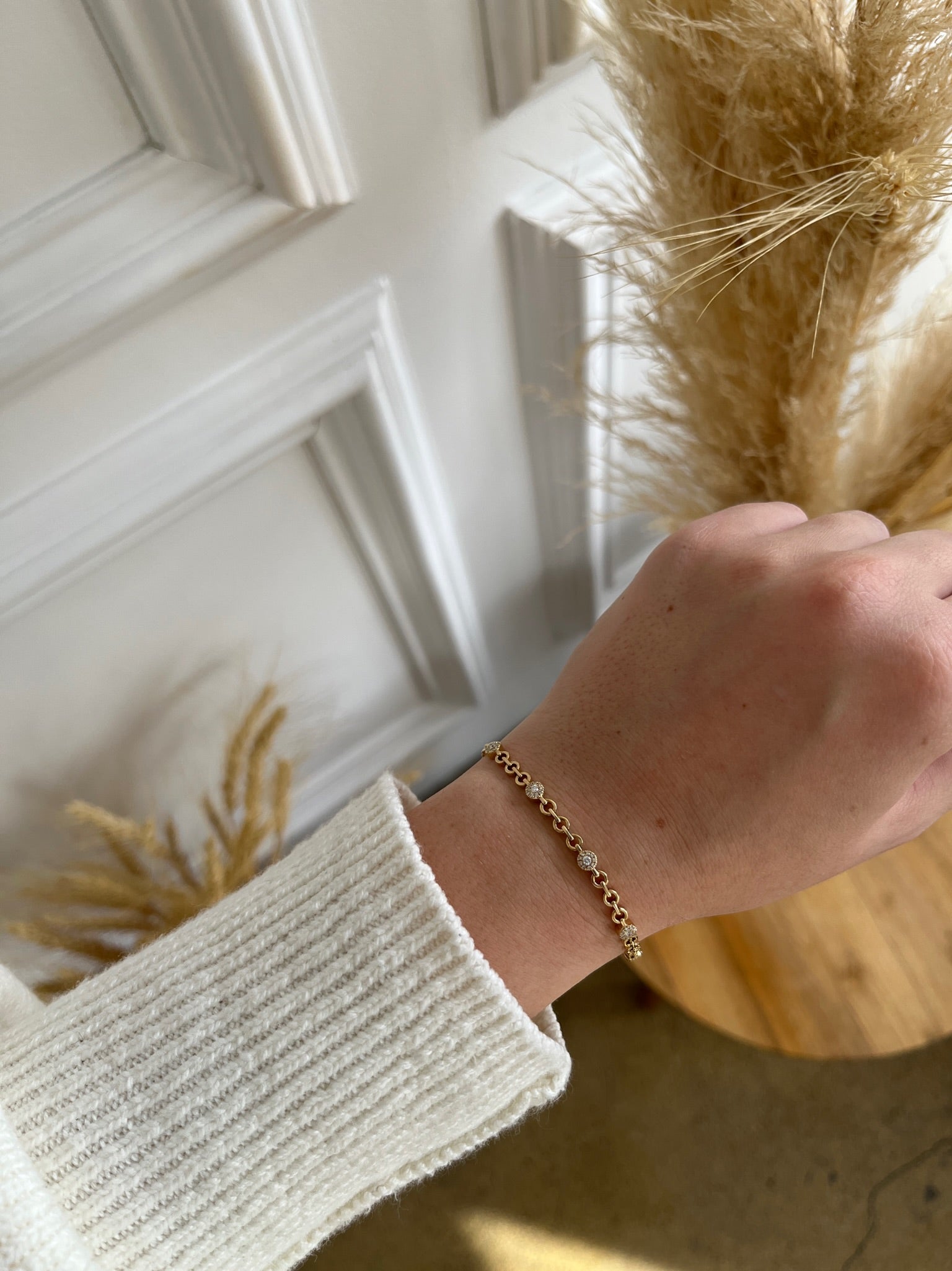 A hand adorned with the Diamond Round Link Bracelet is highlighted as an ideal jewelry piece, paired with a cream ribbed sweater. A white paneled wall and dried pampas grass on wood complete the background.