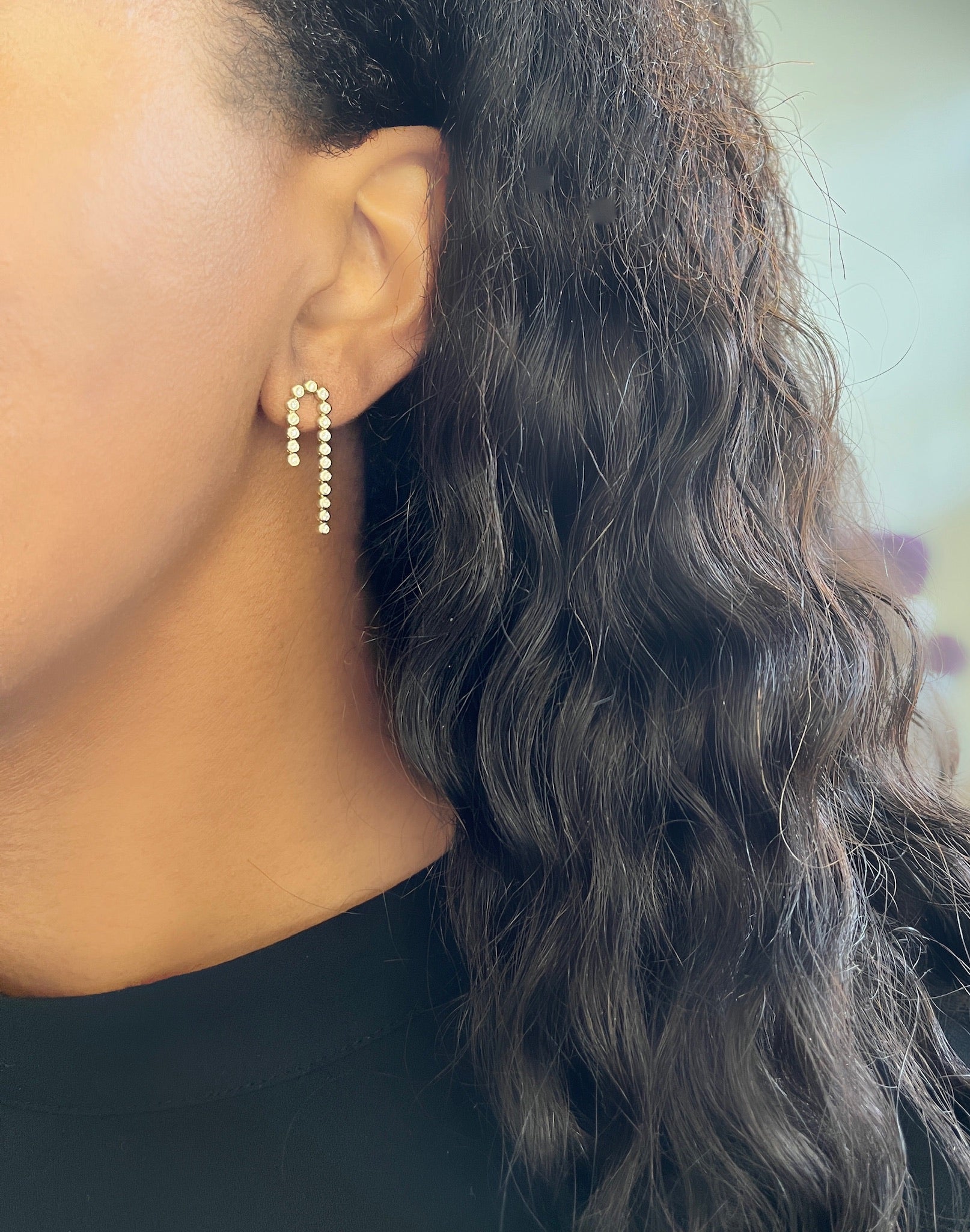 A person with long, wavy black hair is wearing Janet Heller Fine Jewelrys Diamond Waterfall Drop Studs, featuring small round stones in a U-shape. The earrings beautifully complement the black top against a soft blur of pale colors.