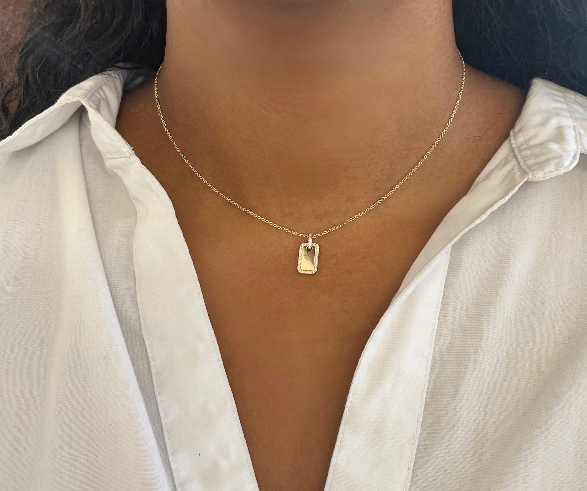 A person wearing a white collared shirt and the Engravable Diamond Dog Tag Necklace—featuring a delicate gold chain and a small rectangular pendant perfect for custom engravings—beautifully showcases the elegance of personalized jewelry.