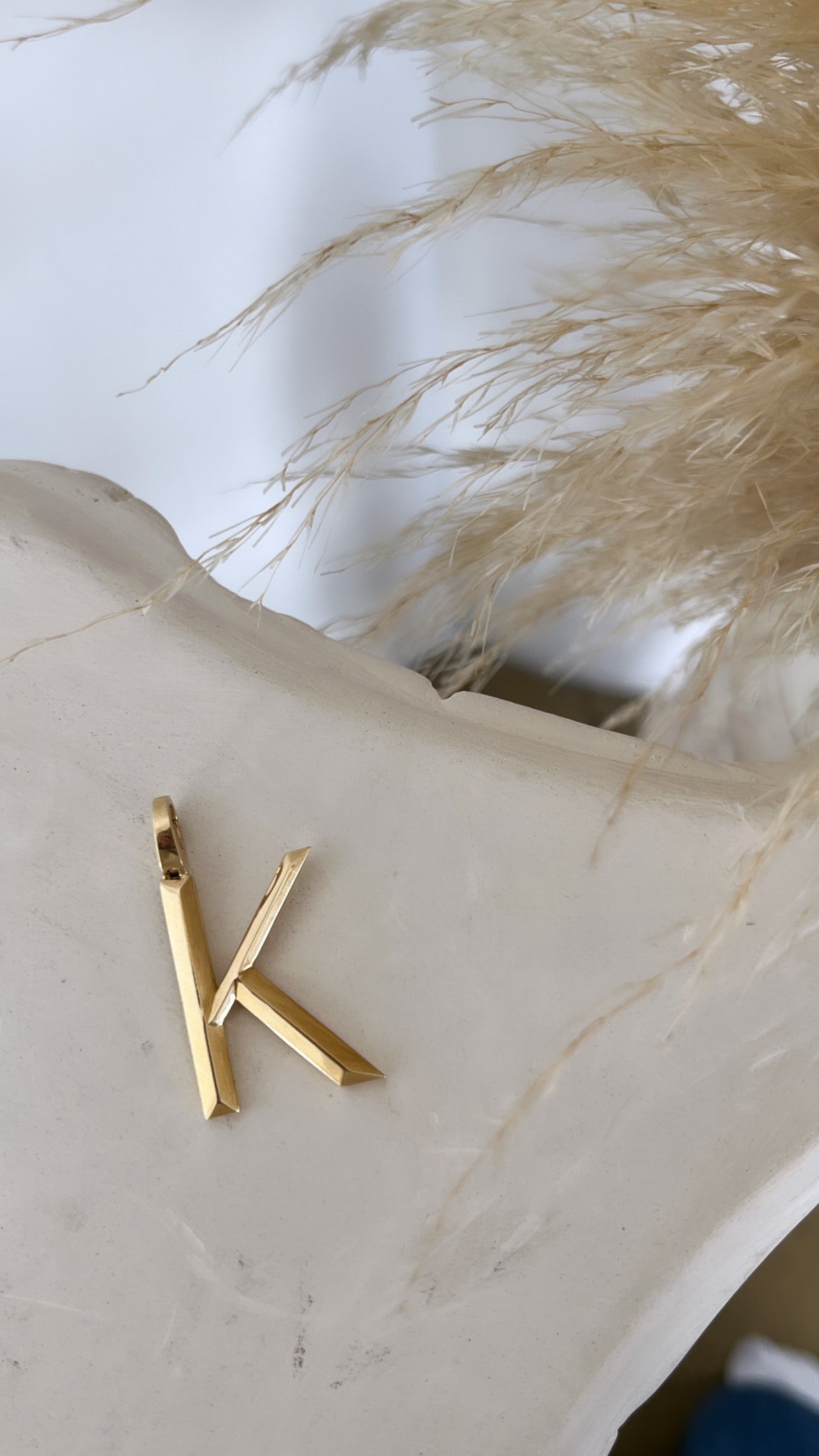The Large Pyramid Initial Charm from FoundryFineJewelry gleams on a smooth, light surface with dried pampas grass nearby. It exudes elegance and simplicity, ideal for pairing with dainty chains or an intricate clasp for extra charm.