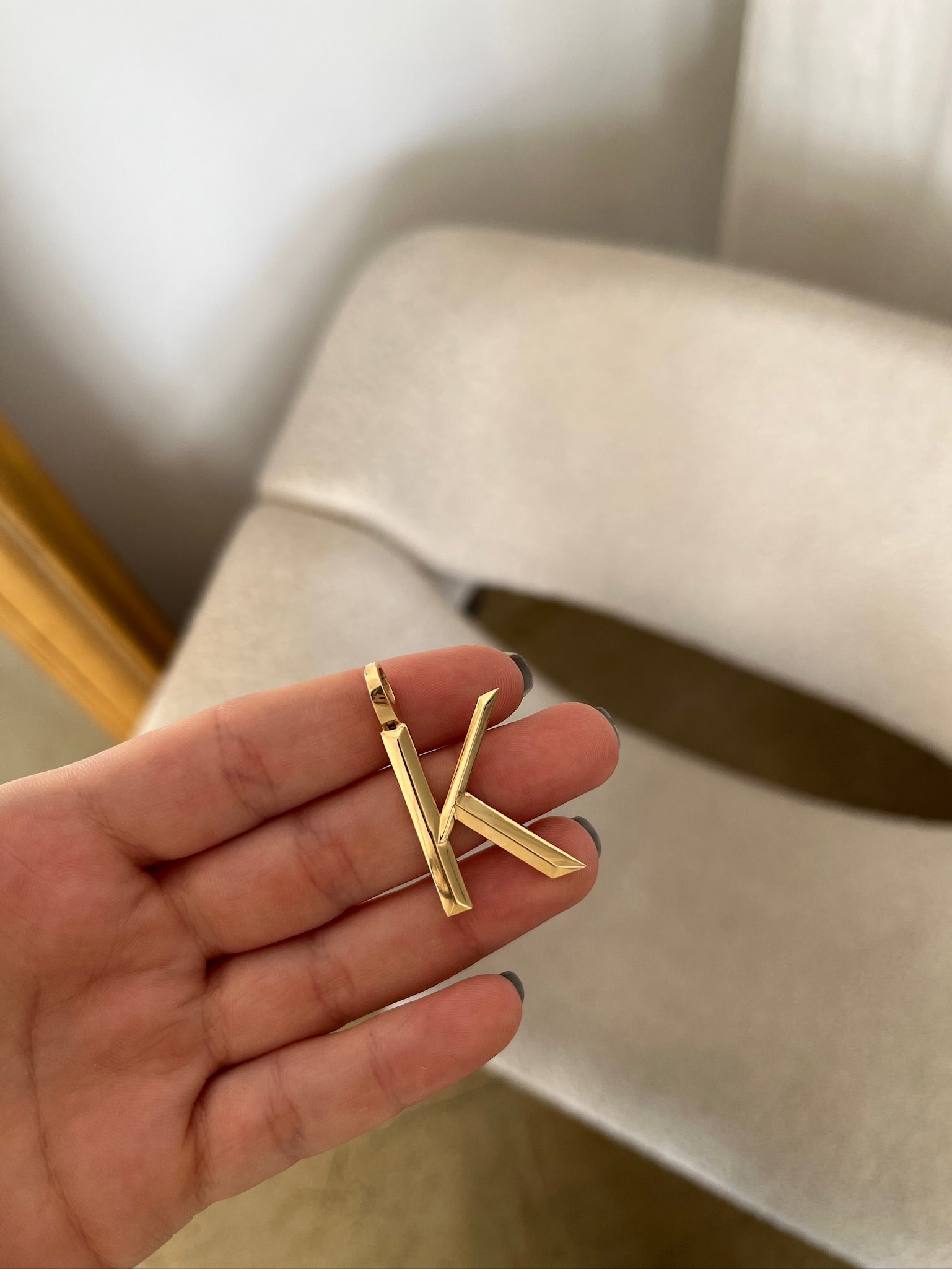 A hand holds the FoundryFineJewelrys Large Pyramid Initial Charm, a gold pendant shaped like K, gleaming elegantly against a beige chair in the background.
