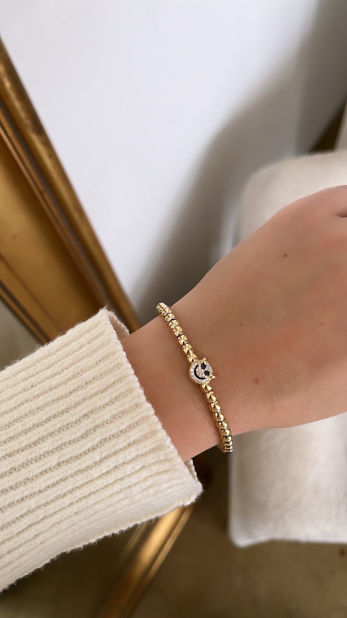 A wrist is adorned with the Little Devil Stretchy Bracelet, showcasing an elegant gold design with a diamond-like centerpiece. It elegantly contrasts with a cream sweater sleeve, while a hint of a gold-colored frame adds allure in the background.