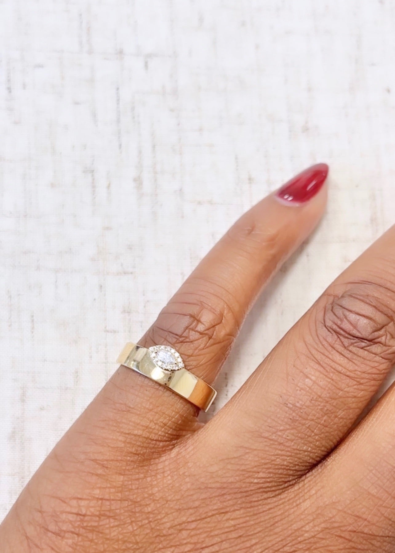 A hand with red polish showcases a classic gold band paired with the Marquise Diamond Gold Band Ring on the ring finger, set against a light, textured backdrop that enhances their allure. These exquisite rings are available for preorder.