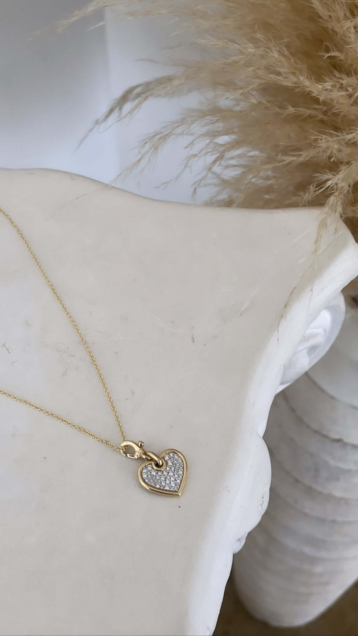 The Medium Flat Diamond Heart Charm from Janet Heller Fine Jewelry, a gold heart-shaped pendant adorned with small diamonds, elegantly rests on smooth white marble. In the backdrop, soft beige pampas grass plumes enhance the charm.