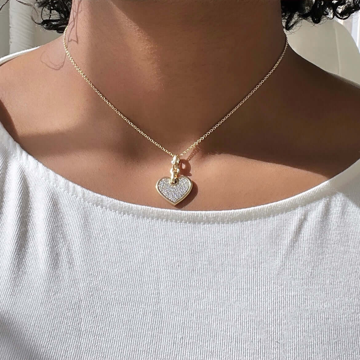 A person in a white shirt wears Janet Heller Fine Jewelrys Medium Flat Diamond Heart Charm, which sparkles alongside a mini bear charm. The softly lit background elegantly highlights the necklaces beauty.