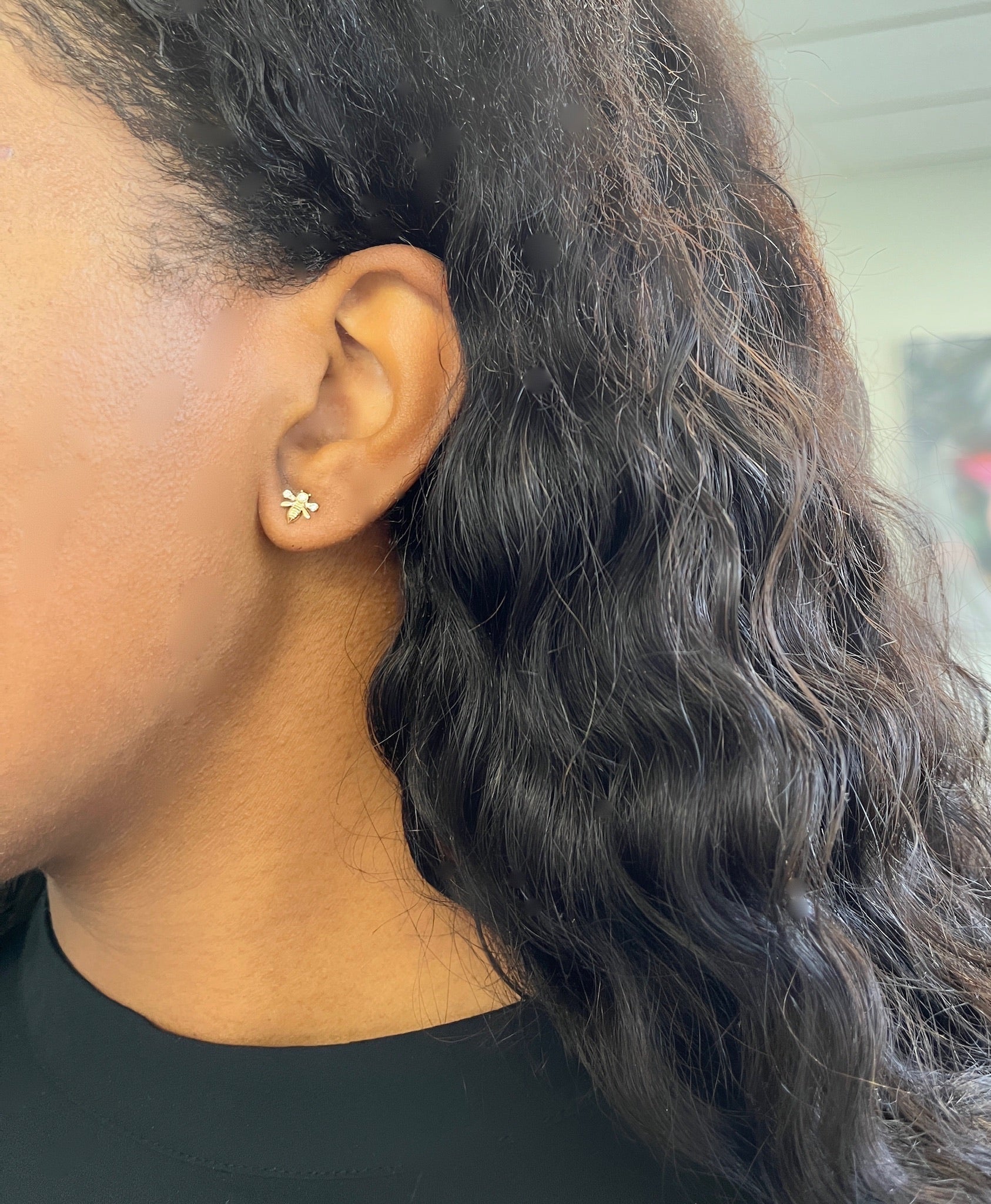 A close-up shows an ear wearing Micro Diamond Bee Studs, framed by long wavy hair that complements a dark top against a softly blurred background, inviting admiration and inquiries about customizations.