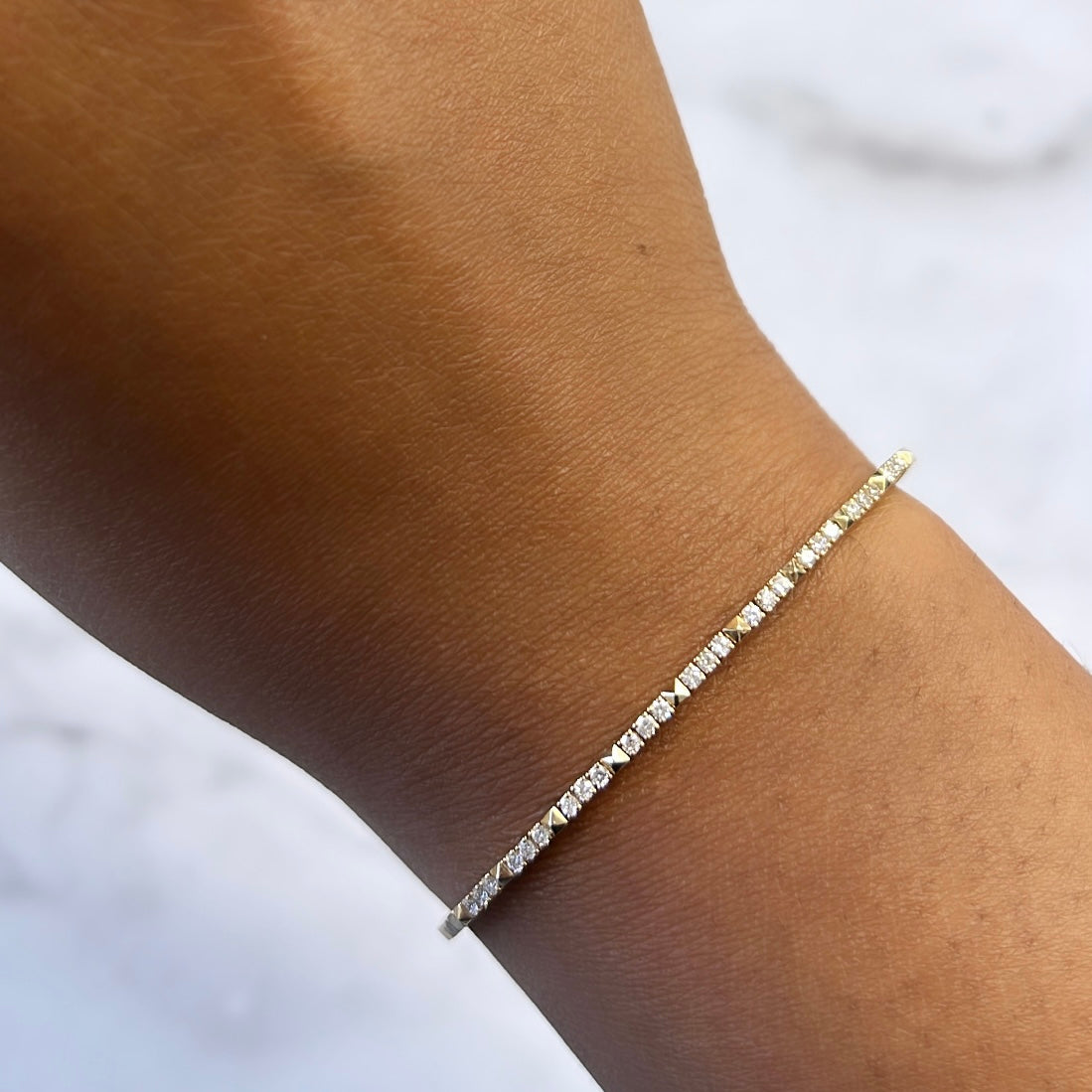 A close-up of a wrist showcases the Janet Heller Micro Diamond Pyramid Bangle, a delicate gold bracelet adorned with sparkling diamonds. The light brown skin pops against the subtle white blur in the background, inviting customizations and inquiries.