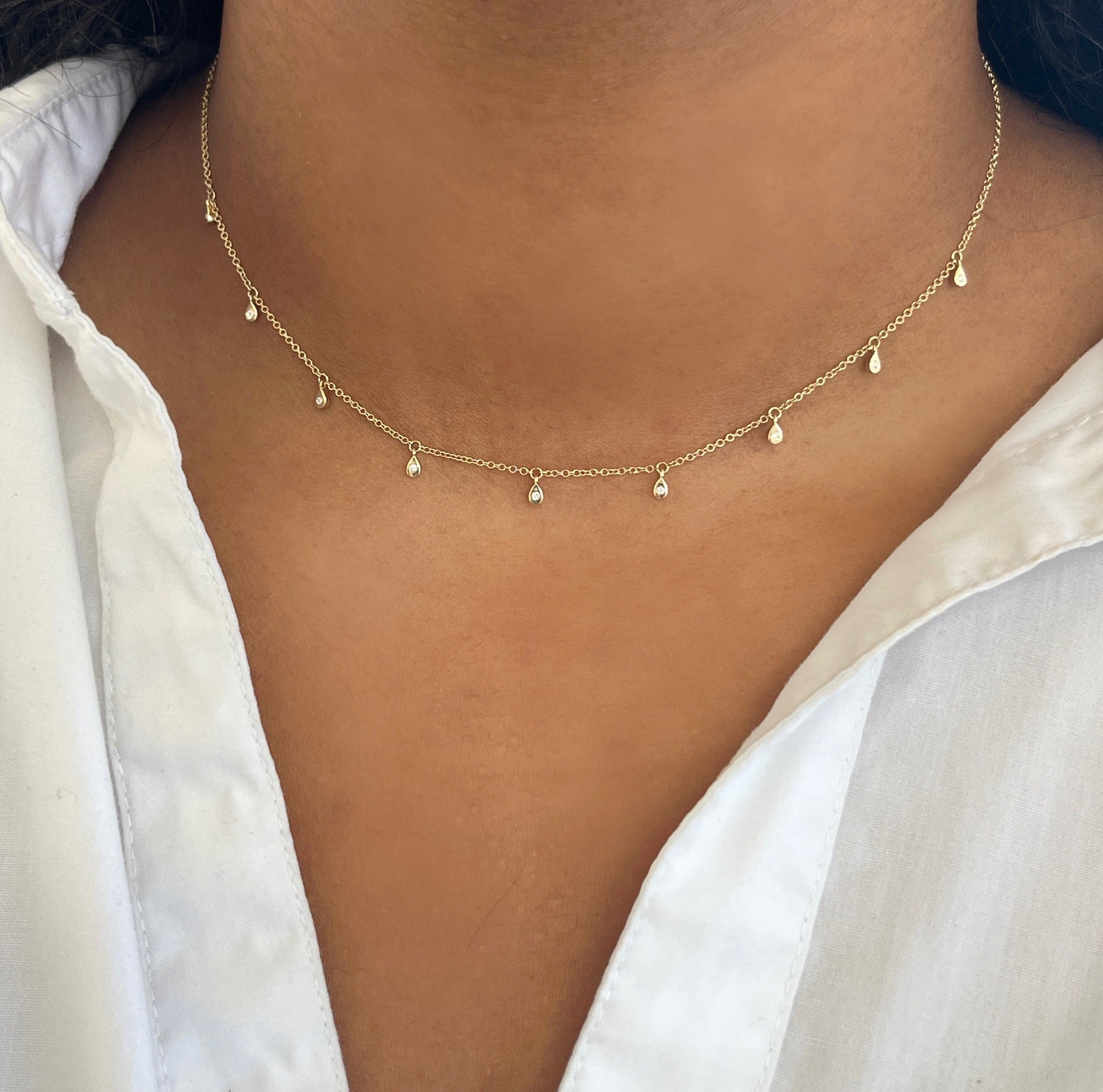 A person elegantly wears the Micro Teardrop Diamond Shaker Necklace, a thin gold piece with dainty charms, perfectly paired with a crisp white collared shirt—a signature item from Janet Heller Fine Jewelry.