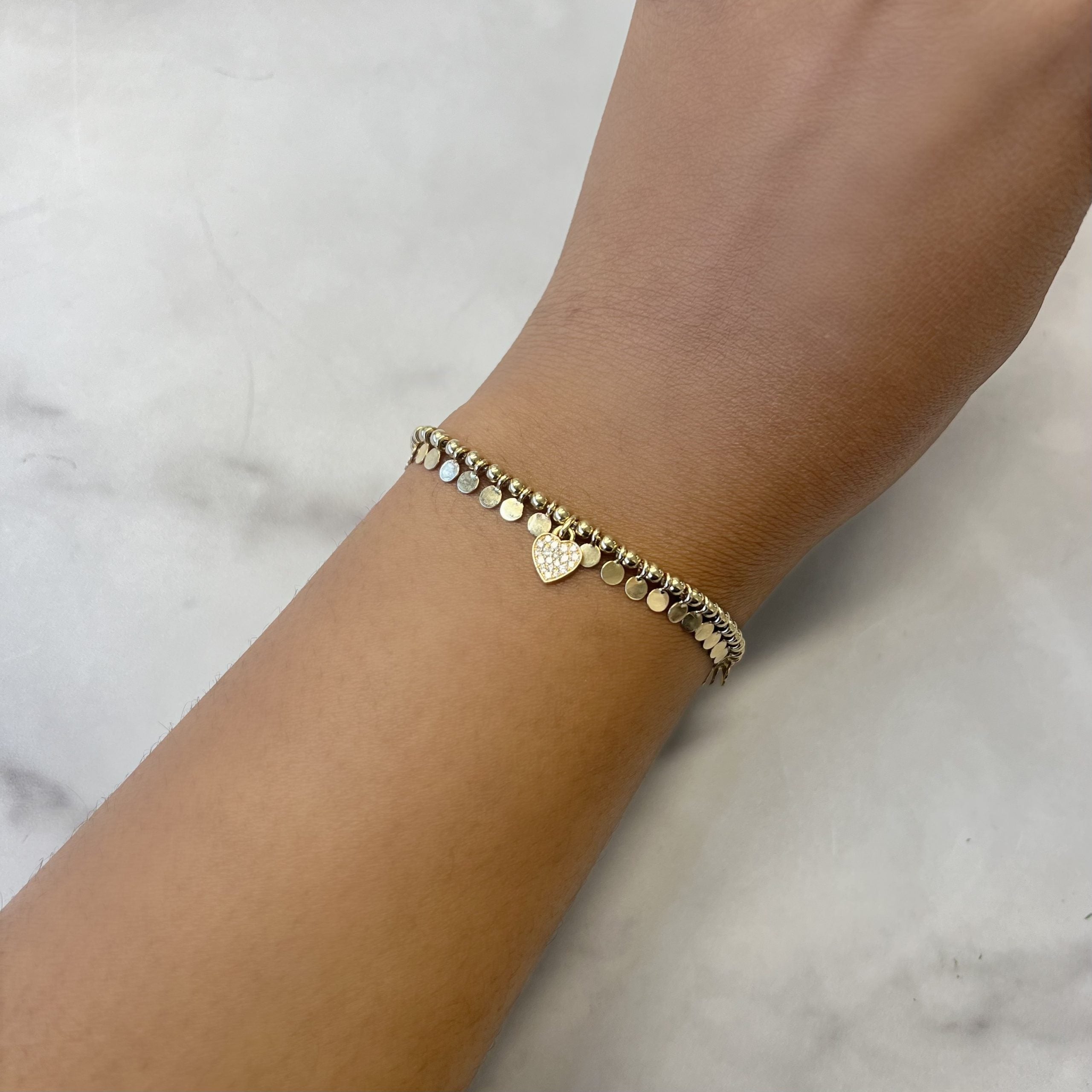 The Mini Coins and Heart Dangle Bracelet from Janet Heller Fine Jewelry elegantly adorns a wrist on a light marble background, featuring small metallic beads and a heart charm with endless customization options for a personal touch.