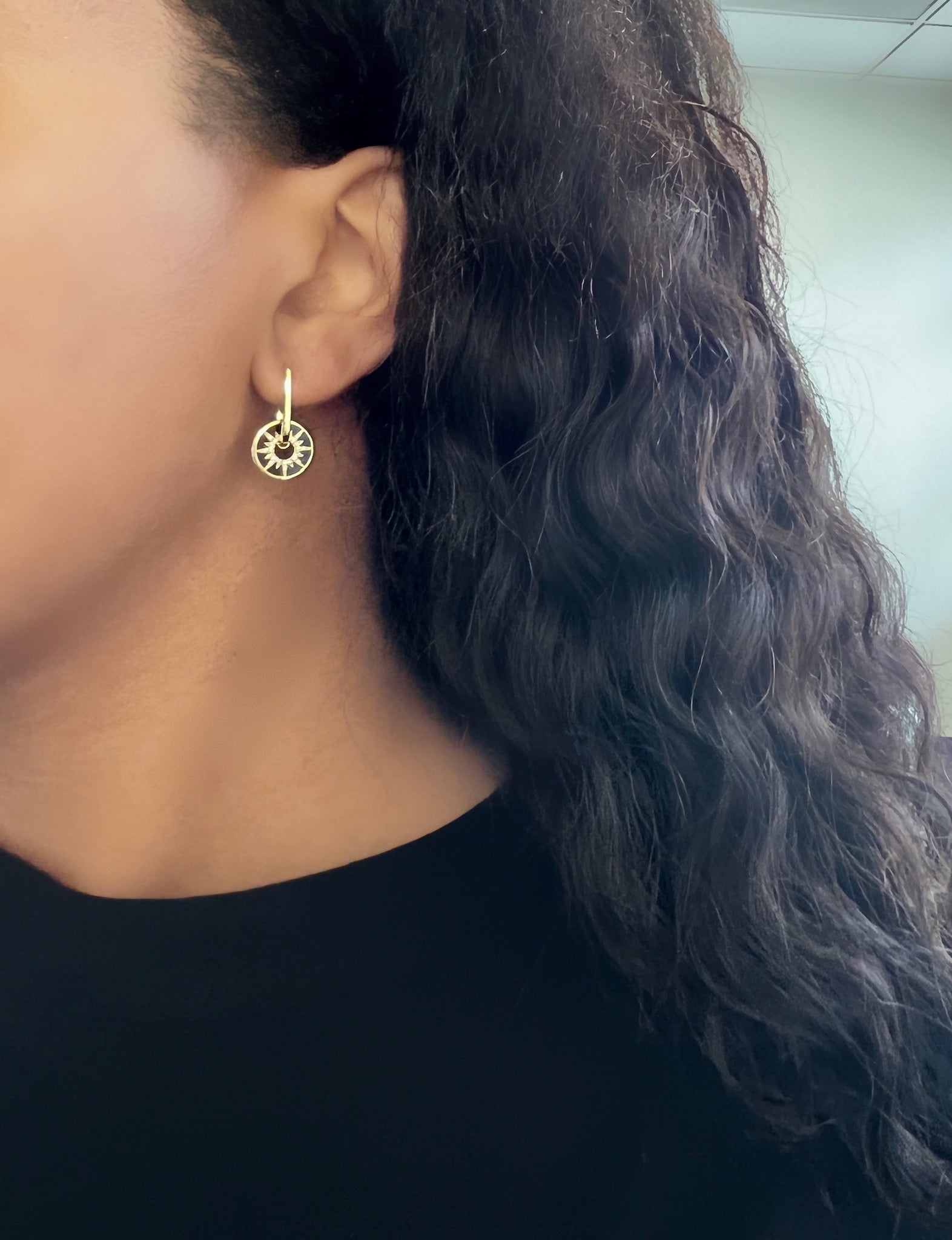 A person with long, wavy hair in a black shirt is shown in close-up profile, wearing Onyx & Diamond Charm Hoops shaped like trees. The softly lit background highlights the artistry of Janet Heller Fine Jewelry.