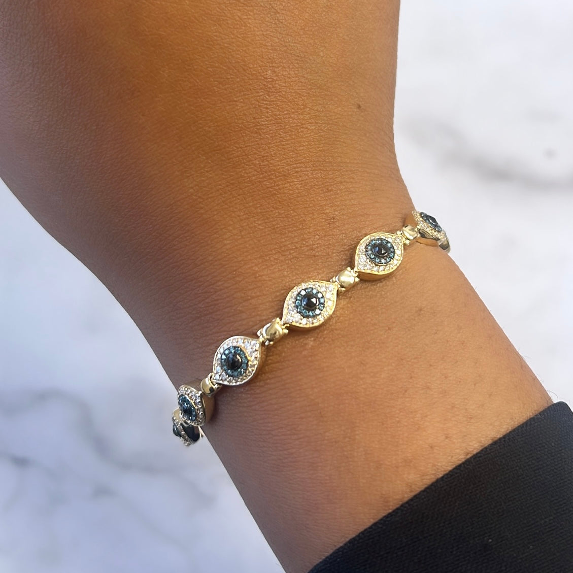 A person wearing the Repeating Evil Eye Bracelet from Janet Heller Fine Jewelry, displaying eye-shaped designs with blue centers and intricate detailing. This exquisite piece on a light background offers customization options for a personalized touch.
