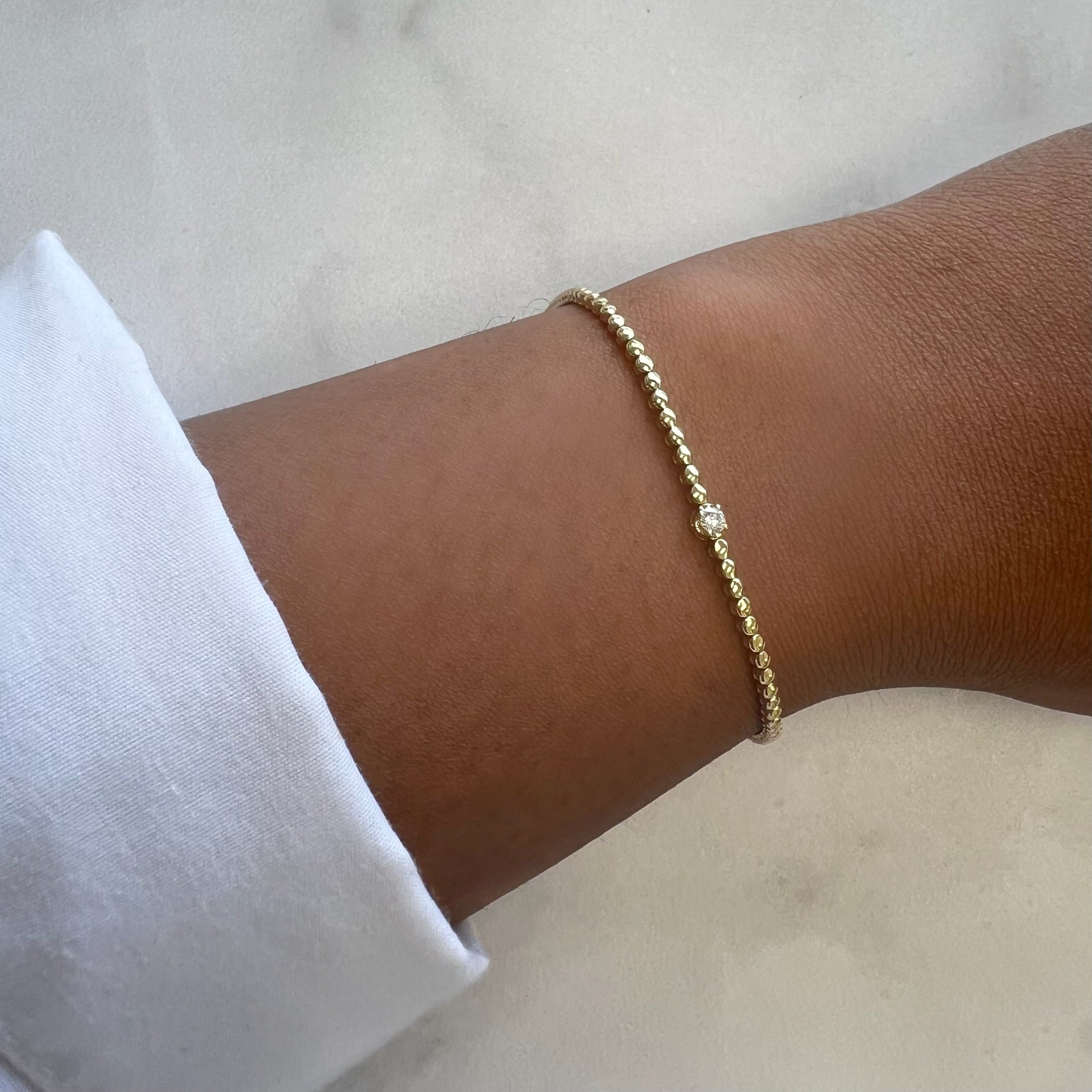 A close-up shows a wrist wearing the Single Diamond Beaded Bracelet from Janet Heller Fine Jewelry. Paired with a white long-sleeve shirt, it stands out against a soft, neutral background.