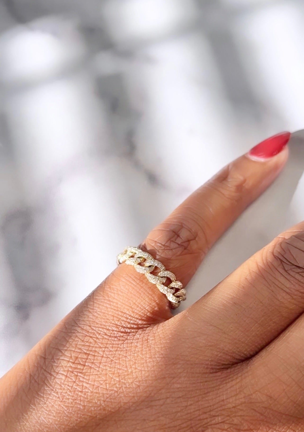 The Small Diamond Chain Ring from Janet Heller Fine Jewelry graces a finger with light brown skin and red nail polish against a soft blur background. This exquisite ring, available for preorder, offers sizing beyond online options.