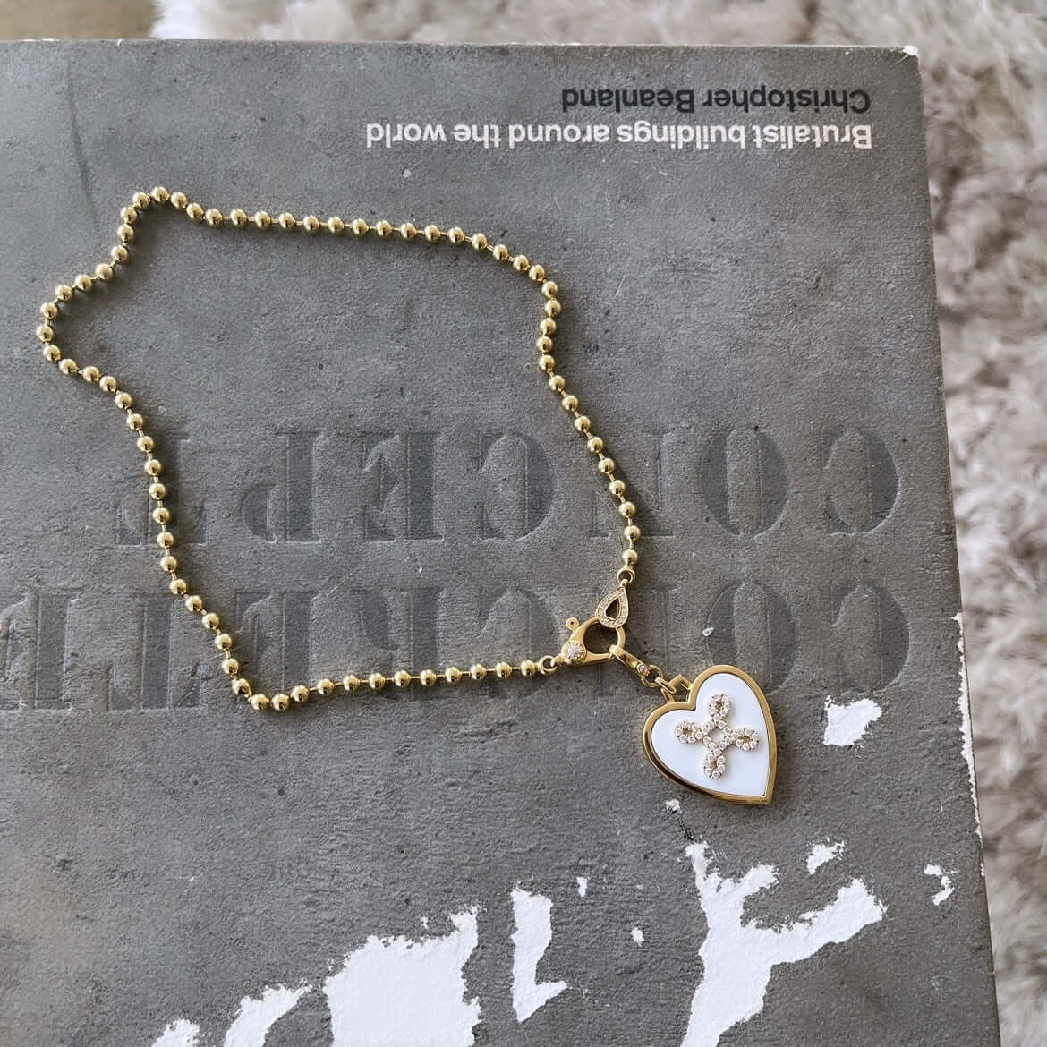 A White Heart Love Knot Charm by Janet Heller Fine Jewelry, featuring a cross design with charms, elegantly rests on a gray book near the corner, showcasing its beautiful clasp.