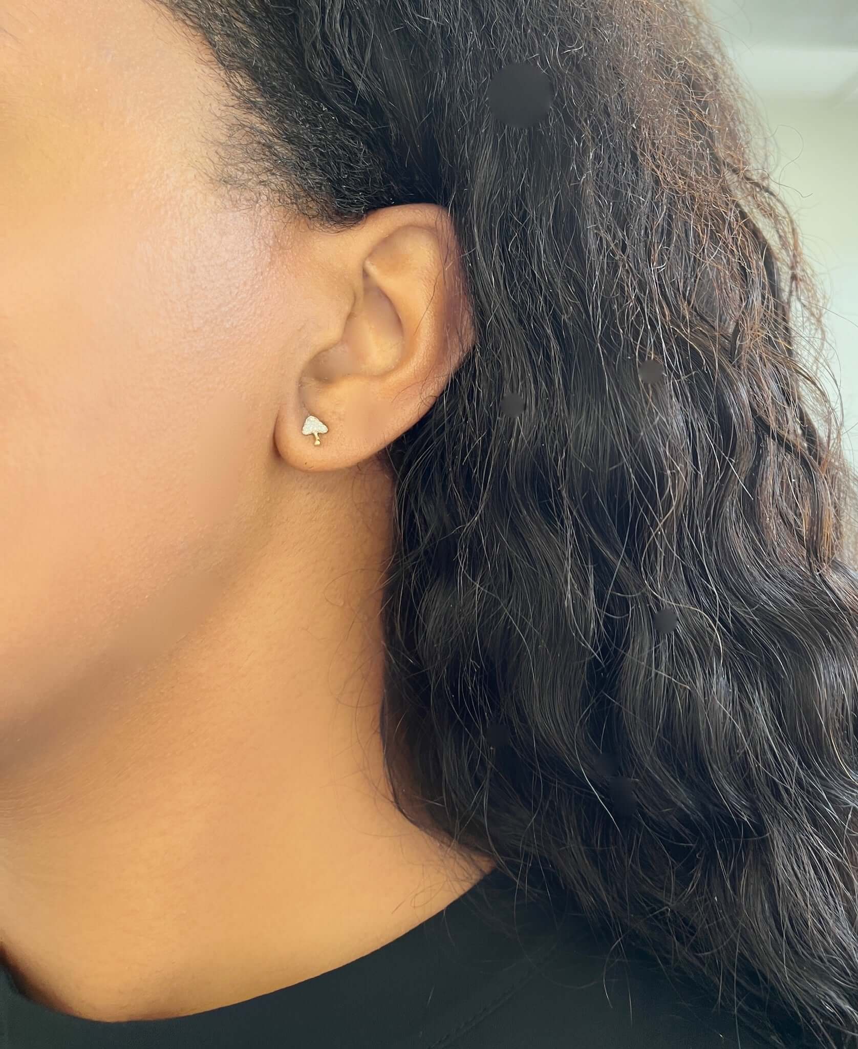 Close-up of a persons ear wearing Diamond Mushroom Studs from Janet Heller Fine Jewelry. The person has long, wavy black hair and wears a black top, with the softly blurred background adding an elegant touch to this exquisite customization.
