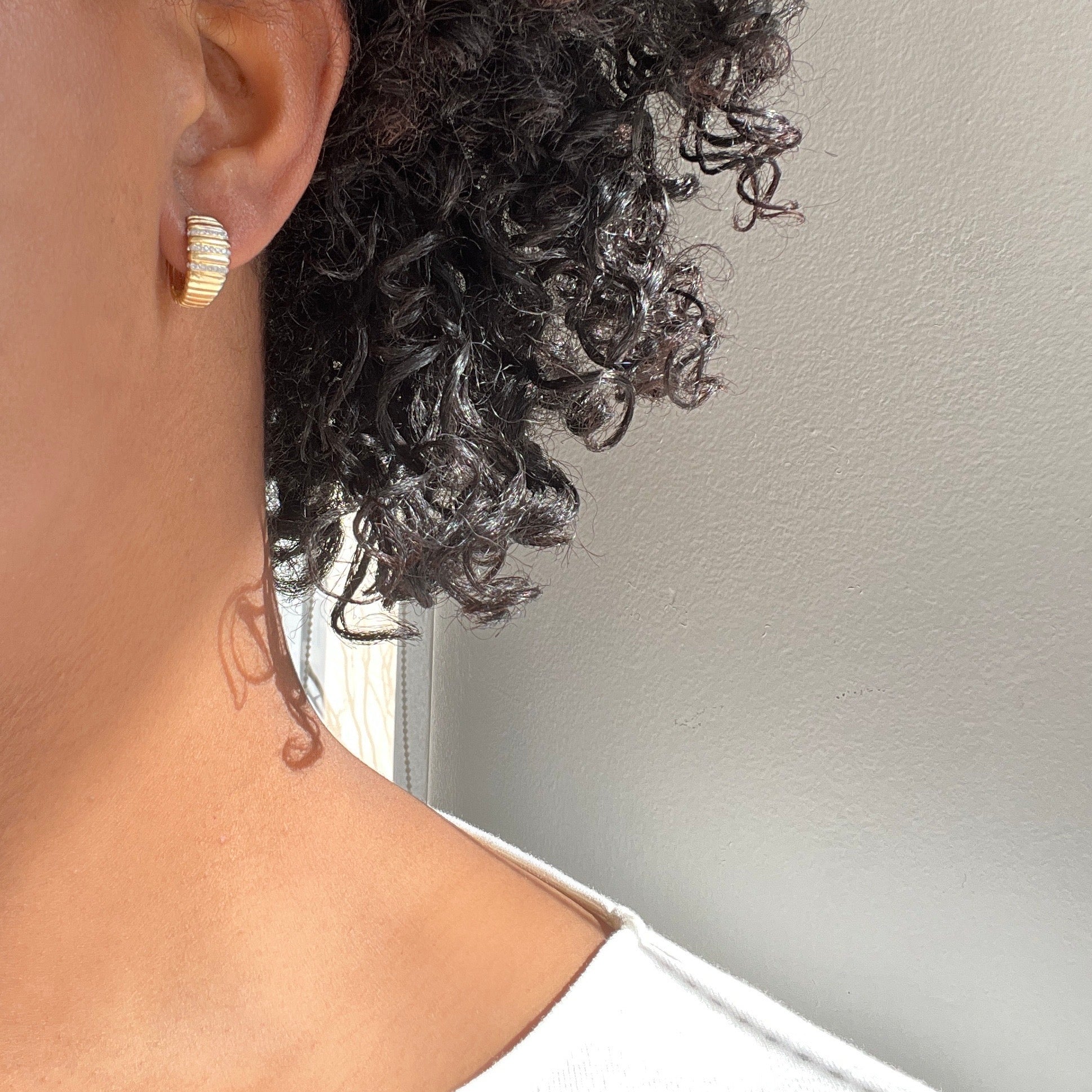 Close-up of a persons neck and ear wearing the Diamond Ridge Huggies by Janet Heller Fine Jewelry. Their short, curly black hair contrasts beautifully against a light gray wall, with a faint shadow adding depth.