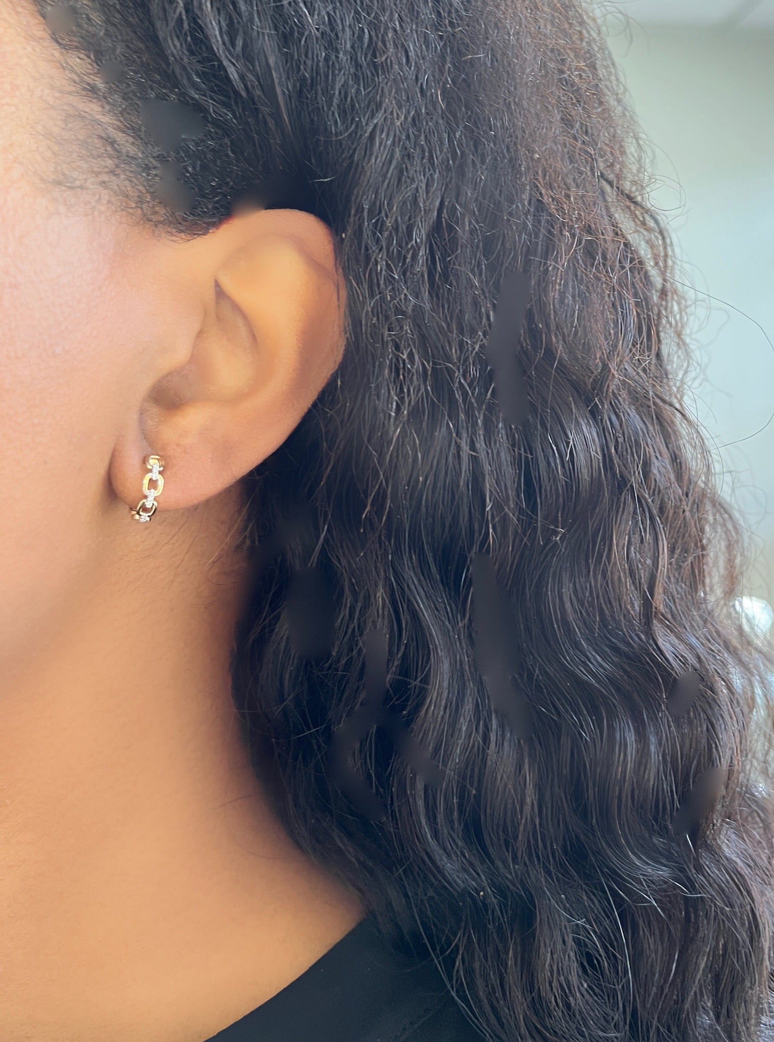 Close-up of a persons left ear with Diamond Square Link Huggies, accompanied by a stud. Their long, wavy black hair drapes down against a blurred, neutral background. For customizations, visit janethellerfinejewelry.com.