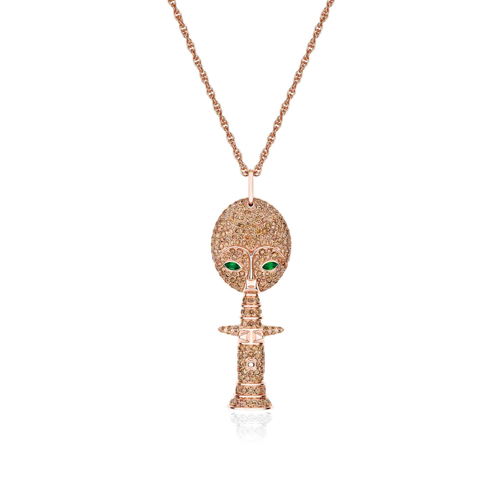 The Brown Mother Moon Goddess Necklace features a gold pendant shaped like a stylized goddess figure, with a round head, green gemstone eyes, and an elongated body with outstretched arms, all embellished with small sparkling stones.