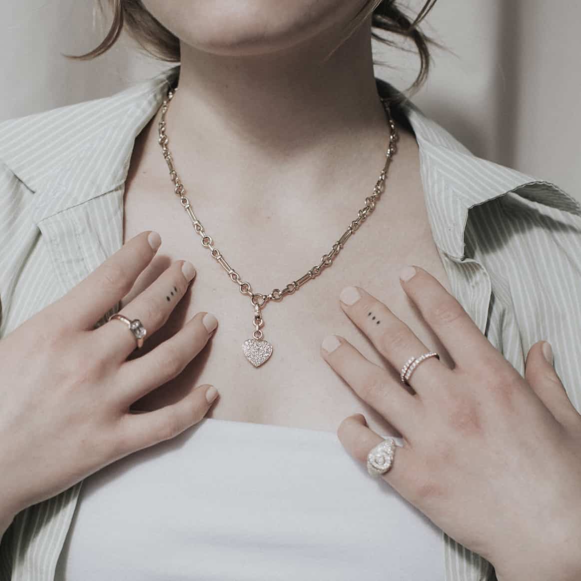 A woman in an open, light-striped shirt and white top displays a gold Lisbon Chain by Janet Heller Fine Jewelry with a heart pendant. Her hands flaunt several rings, charming small tattoos, and nude-colored manicured nails.