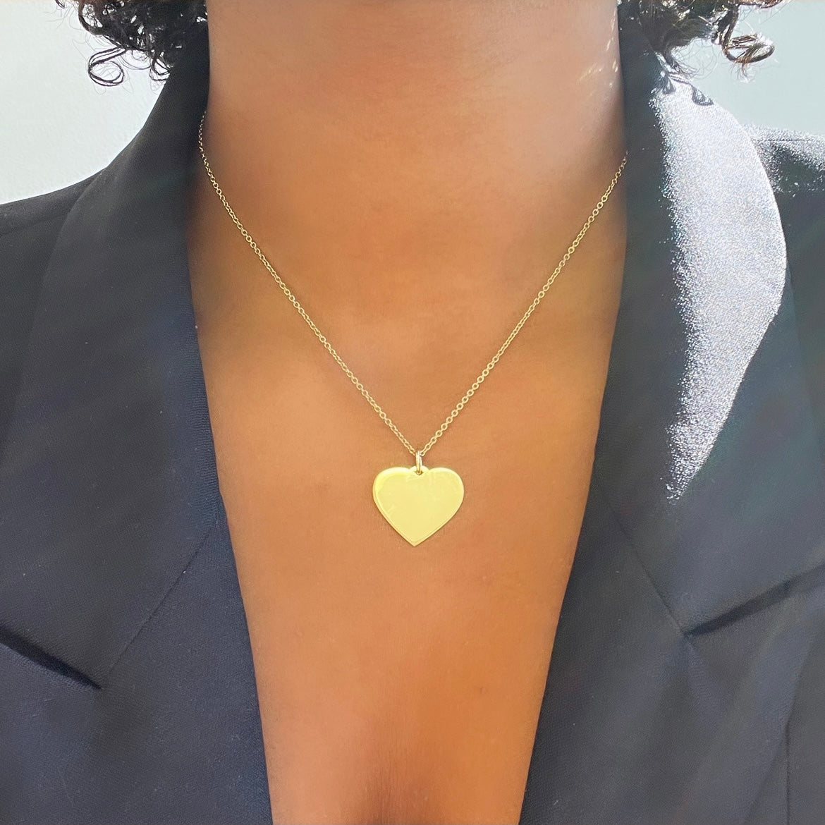 A close-up shows someone in a black blazer wearing a Large Heart Engraveable Pendant Necklace on a delicate chain, with light softly illuminating the elegant jewelry and part of their neck and collarbone.
