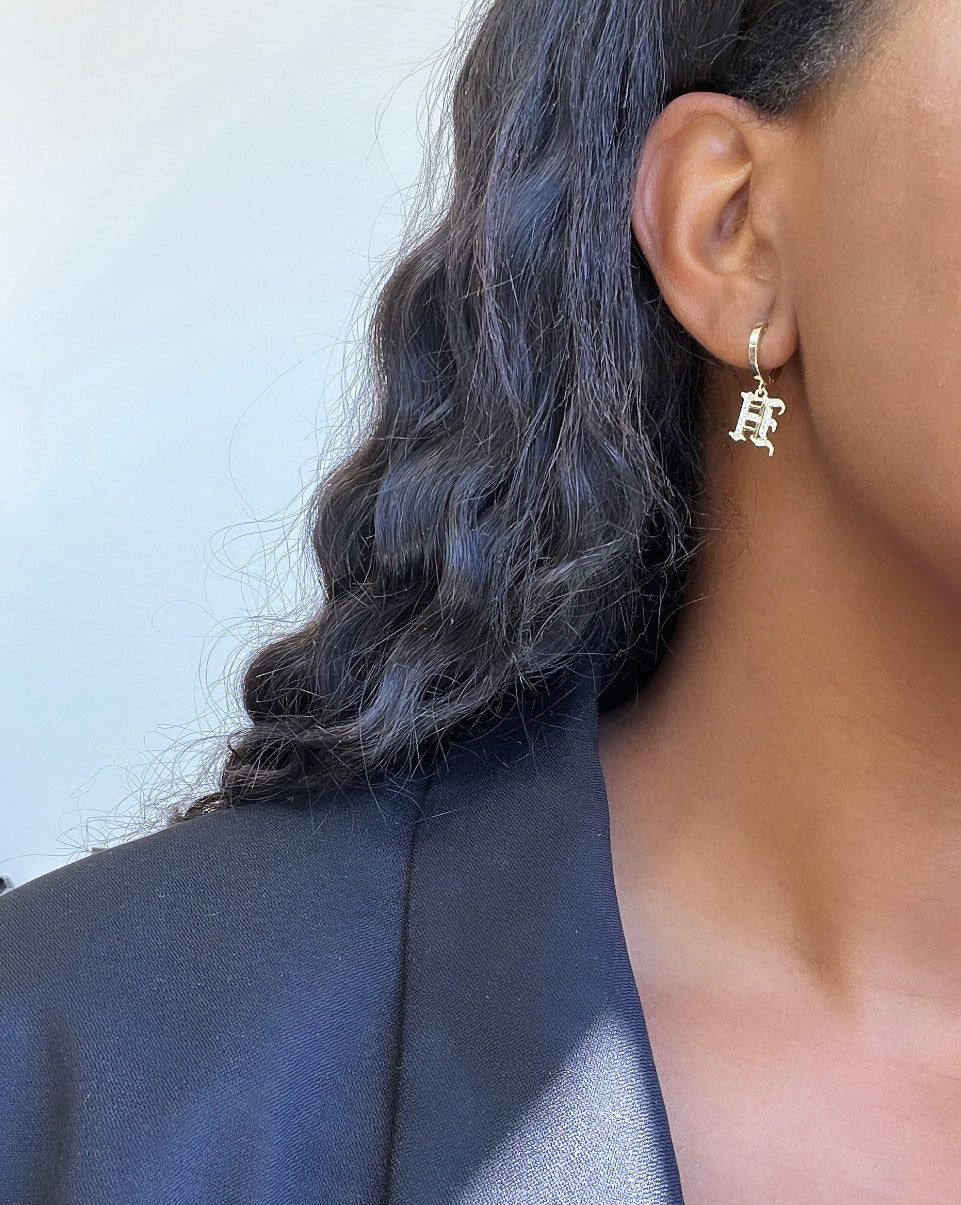 A close-up of a person showcases their distinct style with silver earrings shaped like the letter R, similar to the Small Old English Initial Huggies, complemented by long, wavy black hair and a dark jacket against a blurred background.