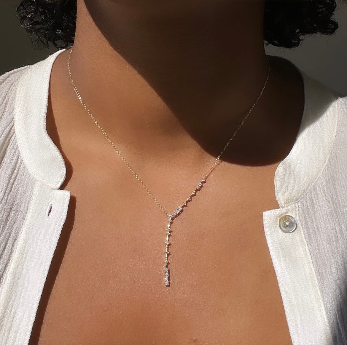 A person wears a white, partially buttoned blouse and a delicate Janet Heller Fine Jewelry Diamond Constellation Lariat Necklace, with small sparkling gems in a vertical line resting on the upper chest.