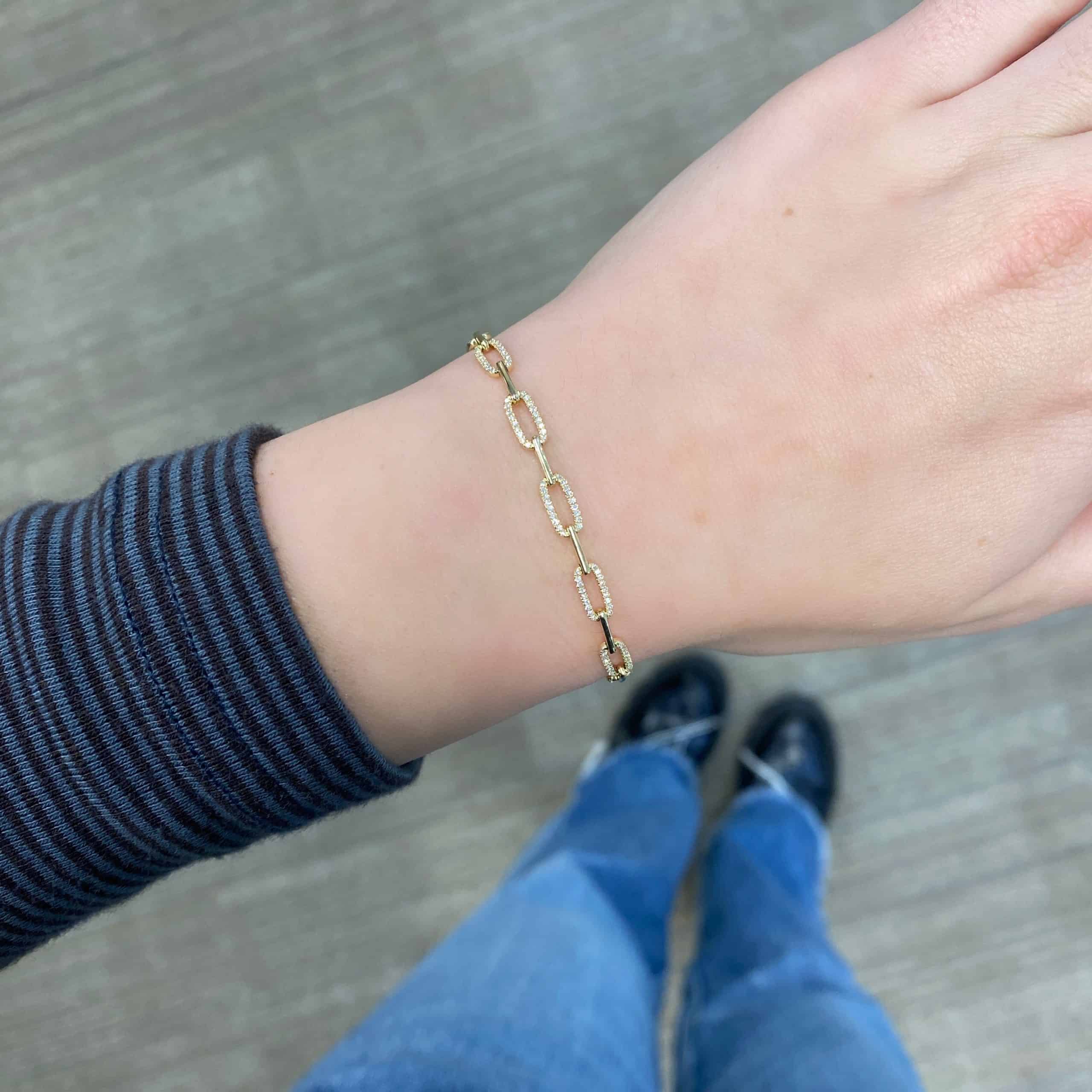 A hand wearing the Alternating Oval Diamond Link Bracelet from Janet Heller Fine Jewelry, with a striped dark sleeve, blue jeans, and black shoes visible against a light-colored floor.