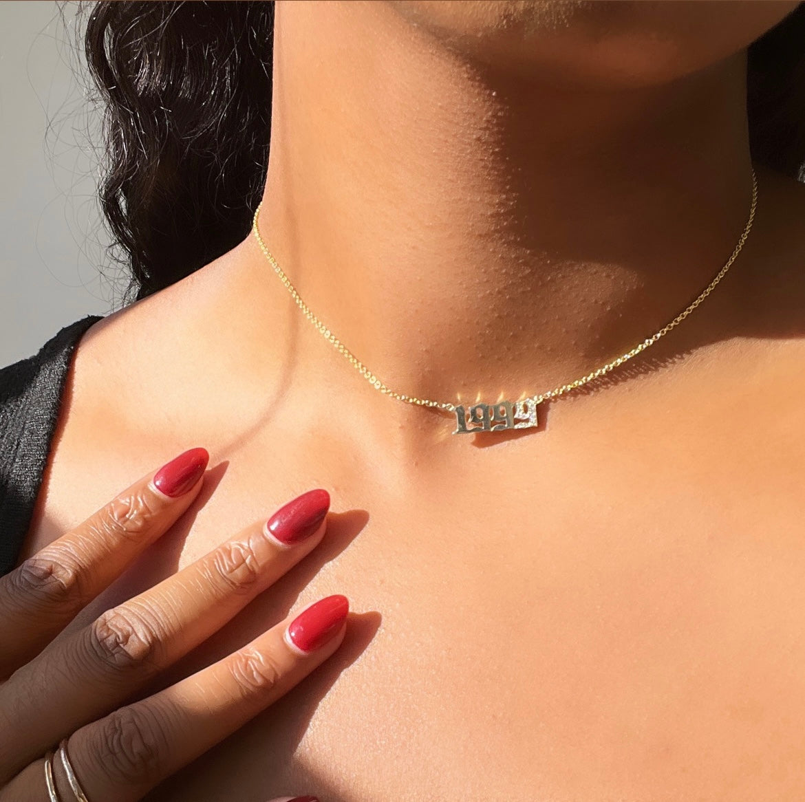 A person showcasing the Diamond Year Necklace, featuring a 1993 pendant, highlights elegant jewelry. Their red nail-polished fingers delicately touch their collarbone as the softly lit background accentuates both the necklaces customizations and their warm skin tones.