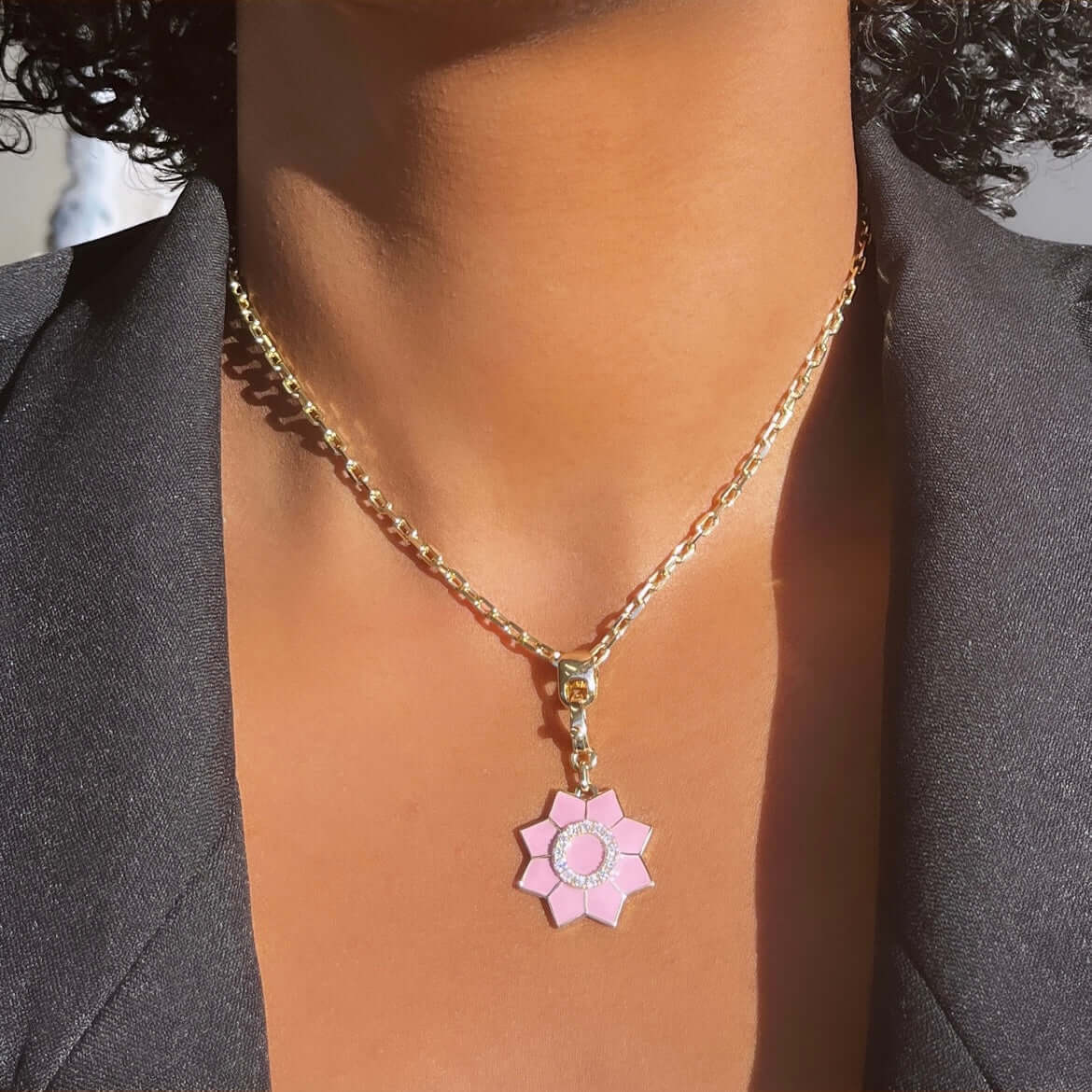 A person in a black blazer wears a Janet Heller Fine Jewelry Pink Enamel & Diamond Flower Charm—a gold chain necklace with a pink flower-shaped pendant, circular center detail, and elegant clasp.