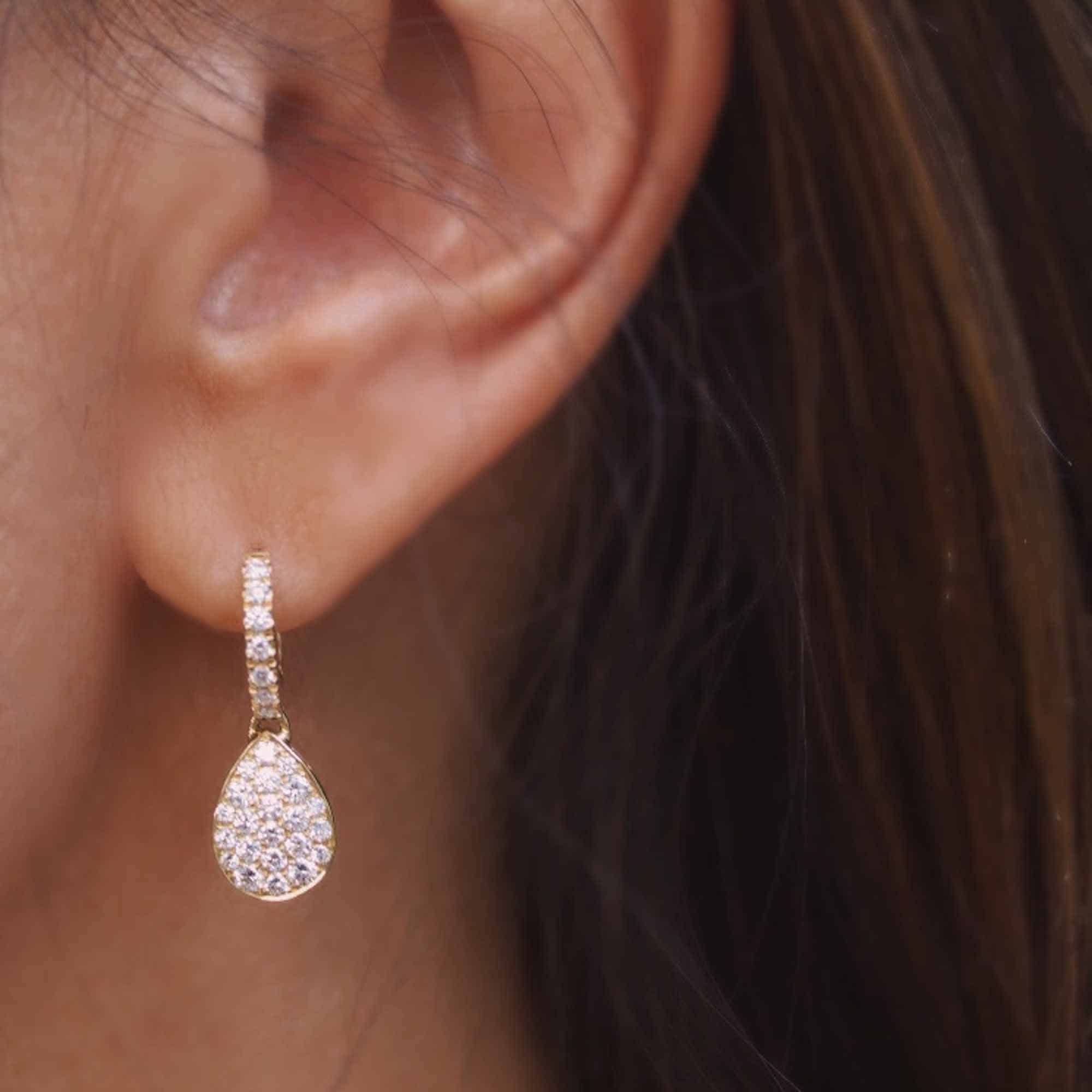 Close-up of a person wearing Janet Heller Fine Jewelrys Pear Shaped Diamond Earrings. The teardrop earring, adorned with sparkling stones, elegantly hangs from a stone-studded hoop. Dark brown hair partially frames the jewelry, enhancing its allure.