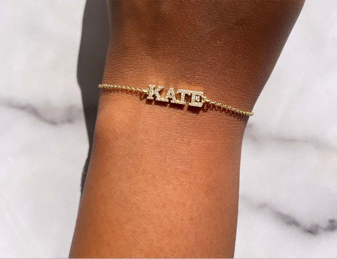 The Diamond Block Name Bracelet is a gold piece with diamond-encrusted letters spelling KATE on a chic marble background, offering a unique way to capture special moments.