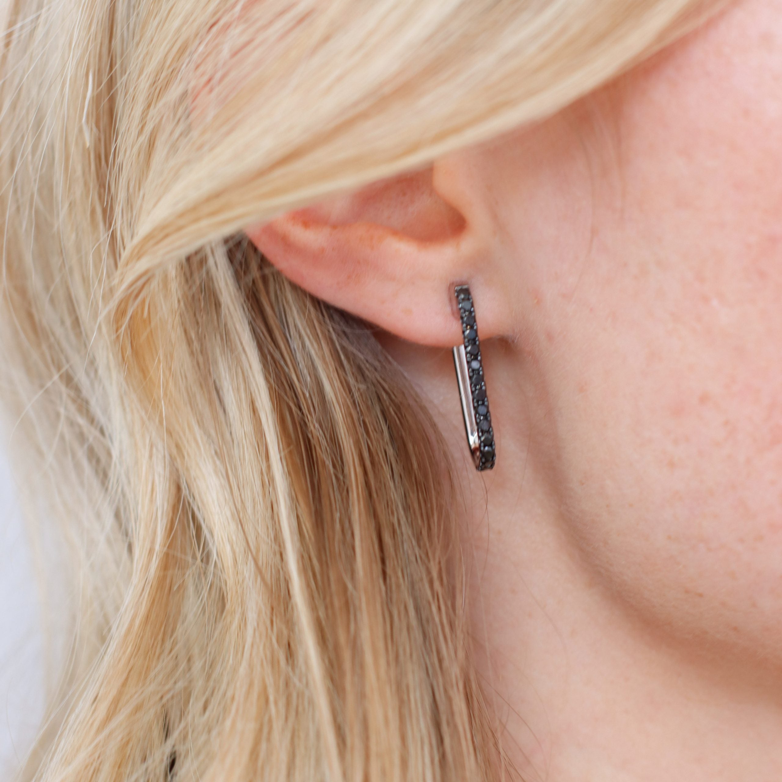 Close-up of a person wearing a Black Diamond Mini Alice Earring on the left ear by Janet Heller Fine Jewelry. The rectangular black earring, with its textured dark surface, complements the light blonde hair and fair skin beautifully.