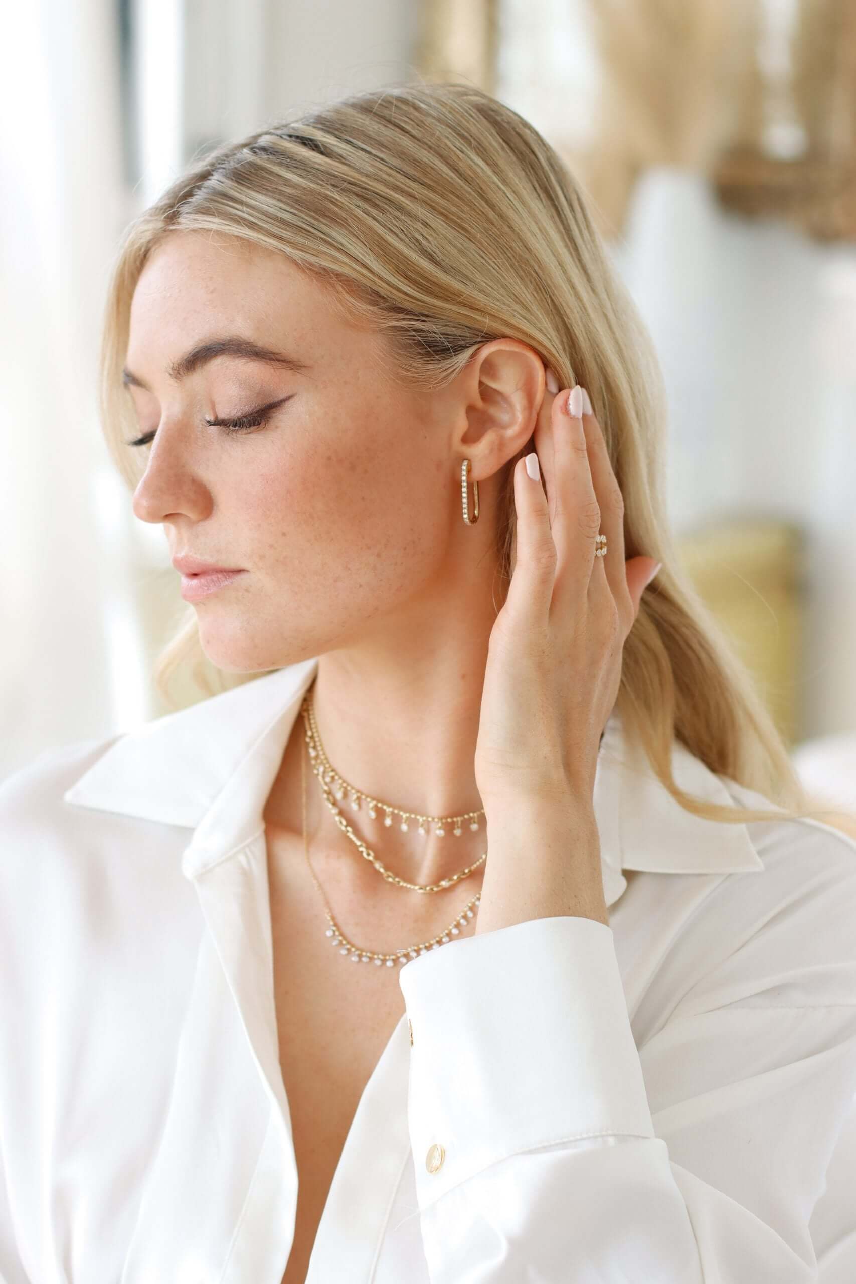 A woman with long blonde hair, in a white blouse and layered gold necklaces, gently touches her ear while wearing Mini Diamond Alice Earrings. Her eyes are closed, highlighting the exquisite jewelry in an indoor setting with soft lighting.