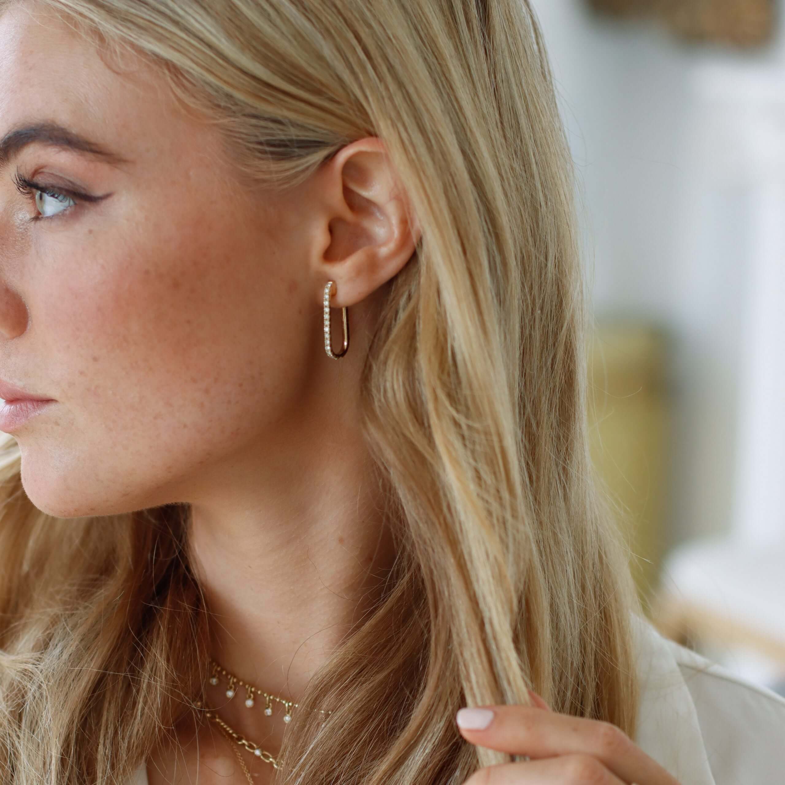 A woman with long blonde hair wears Mini Diamond Alice Earrings and a custom necklace, looking to the side while gently touching her hair. The background is softly blurred.