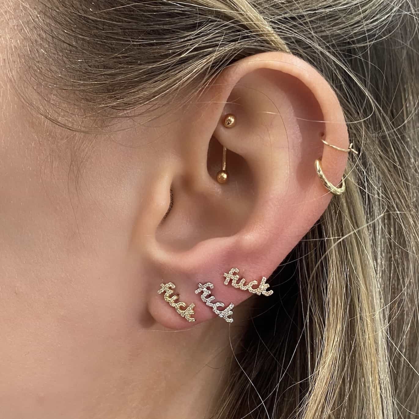 A close-up of an ear with multiple piercings, featuring gold hoops and three Mini Diamond F**k Stud earrings in cursive letters.