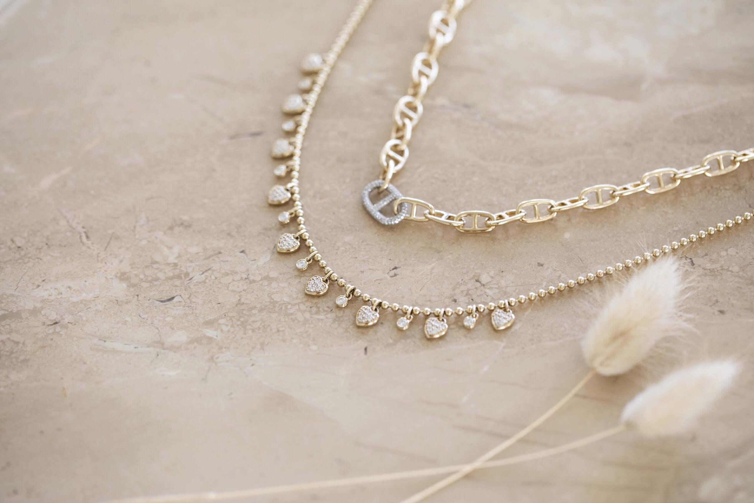 The Diamond Juliette Necklace from Janet Heller Fine Jewelry rests on a beige stone surface beside another gold necklace; one has a linked chain with a circular clasp, and the other features small pendants and delicate beads. Fluffy beige plants in the foreground lend natural elegance to the setup.