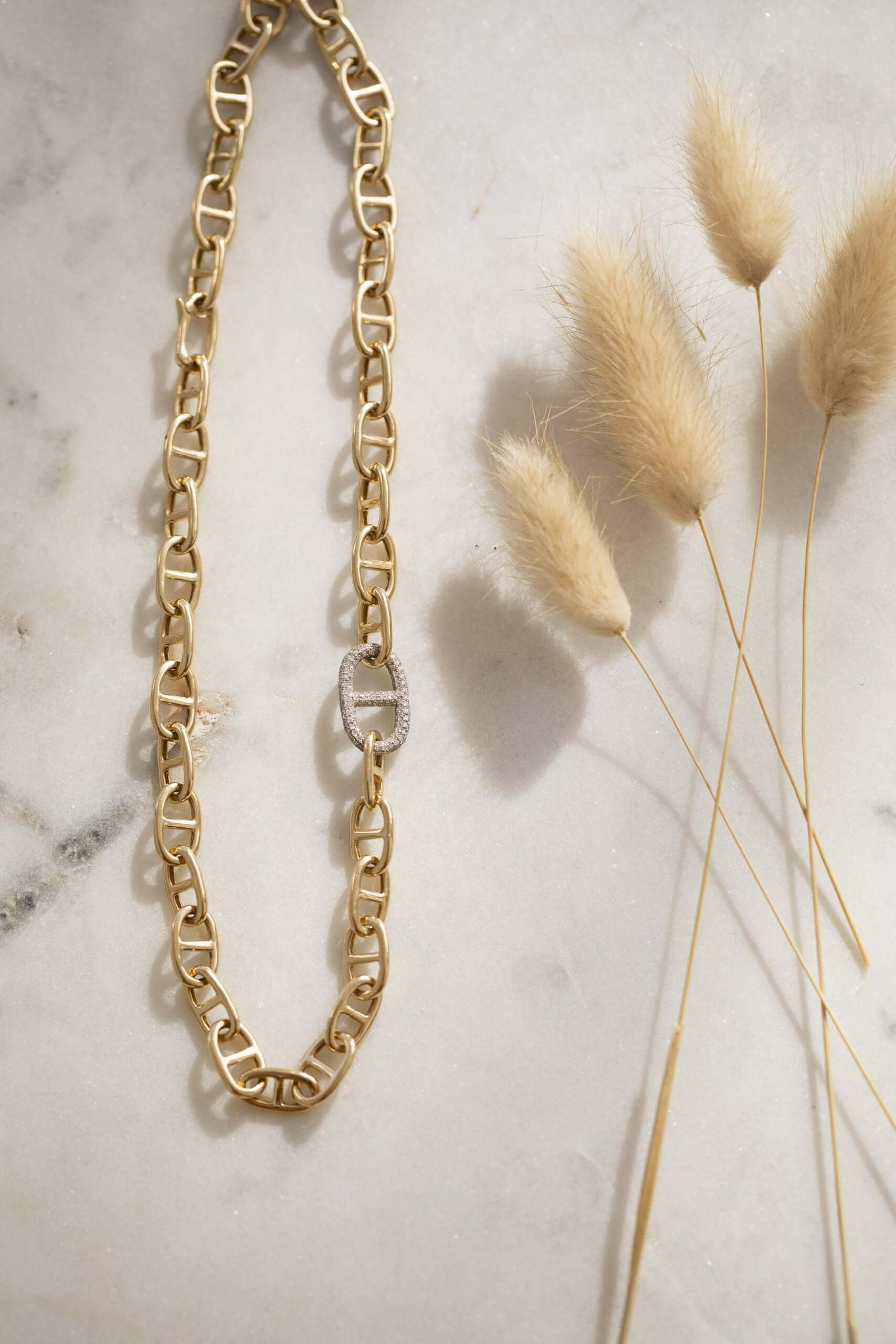 The Diamond Juliette Necklace by Janet Heller Fine Jewelry, featuring a diamond-encrusted link, is elegantly displayed on marble. Delicate dried grass stems nearby add a natural touch, while soft lighting creates gentle shadows, enhancing its presentation.