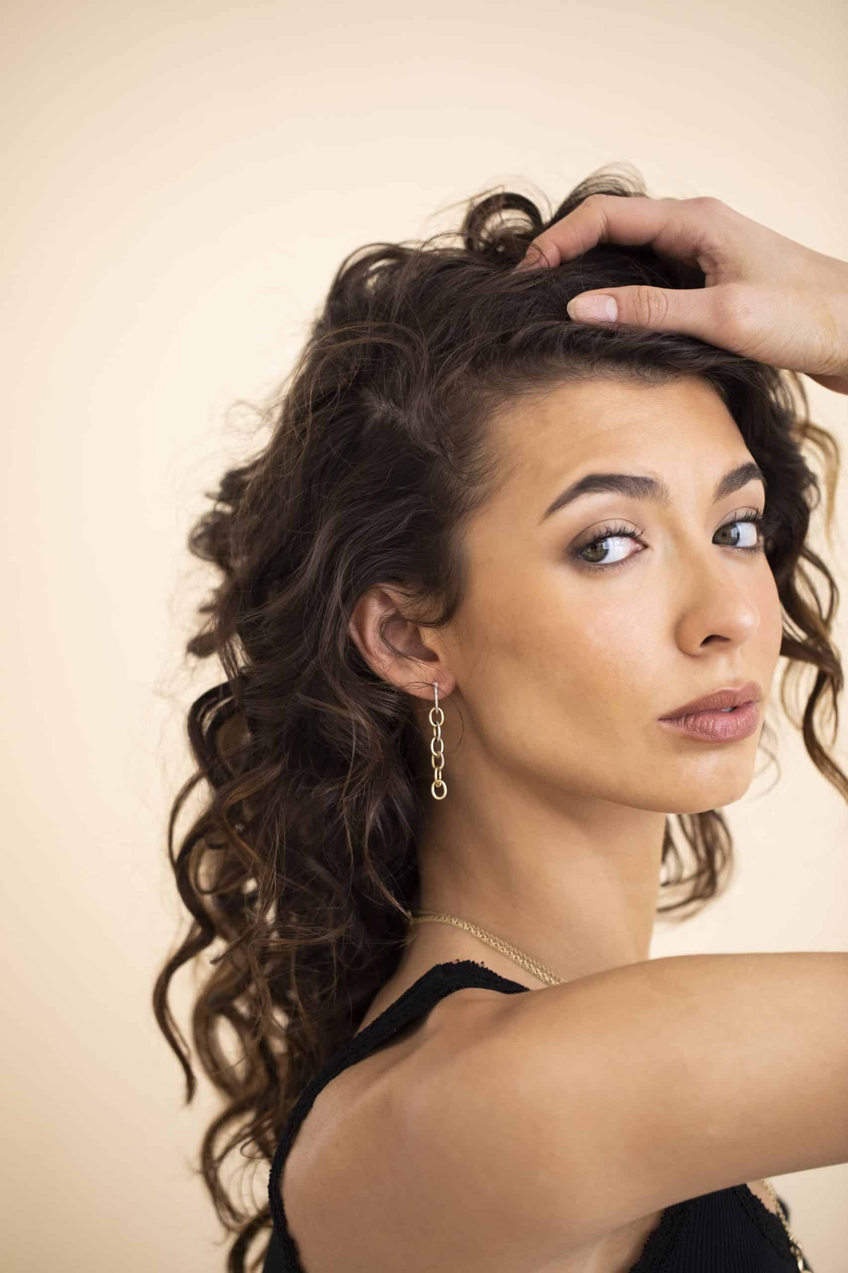 A woman with curly brown hair poses with her hand on her head, wearing a black top and Janet Heller Fine Jewelrys Diamond Round Link Drop Earrings, looking over her shoulder against a neutral background.
