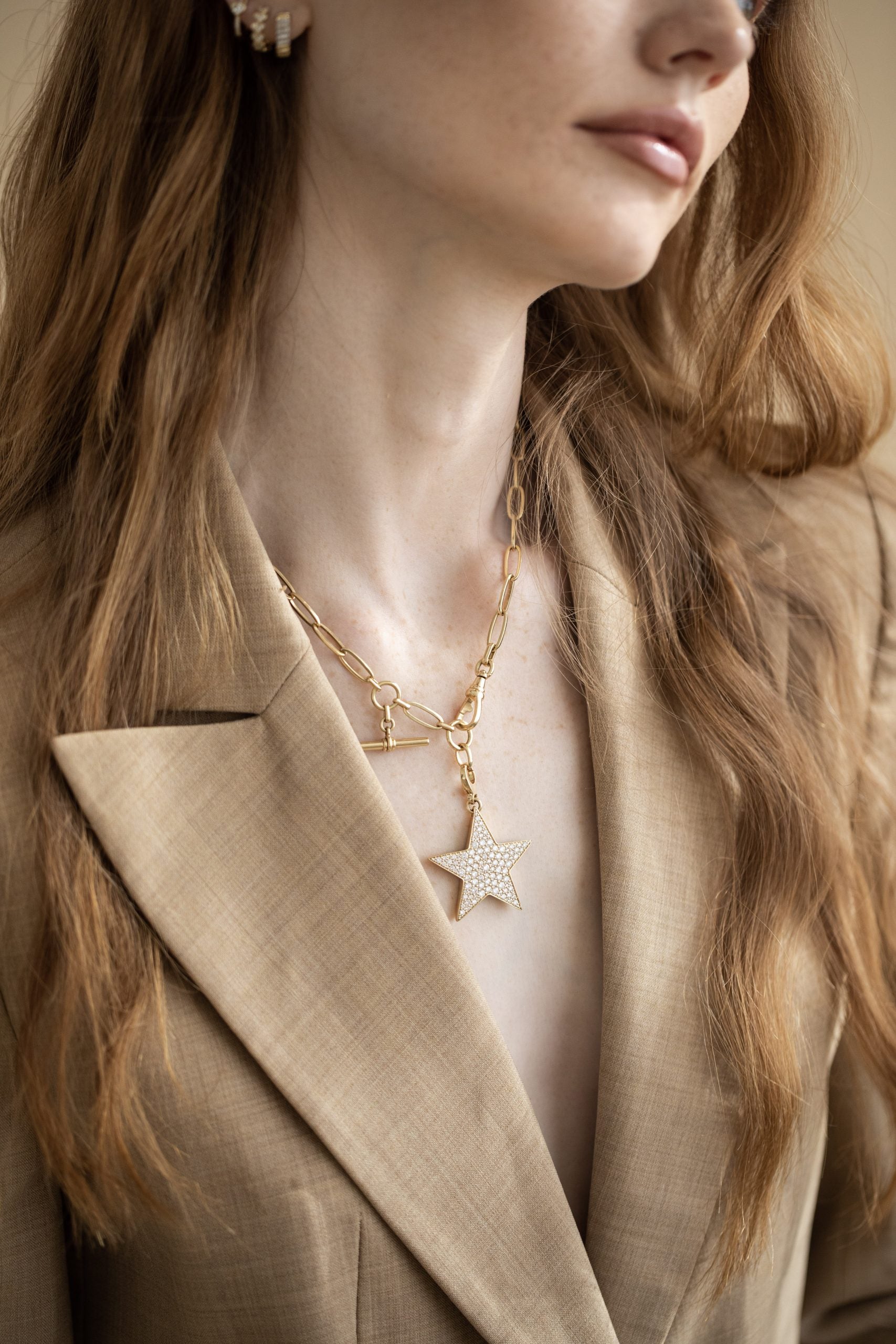 A woman with long, wavy hair wears a tan blazer, accented by the Jumbo Diamond Star Charm from Janet Heller Fine Jewelry on a gold chain necklace and small earrings. Her face is mostly out of view.