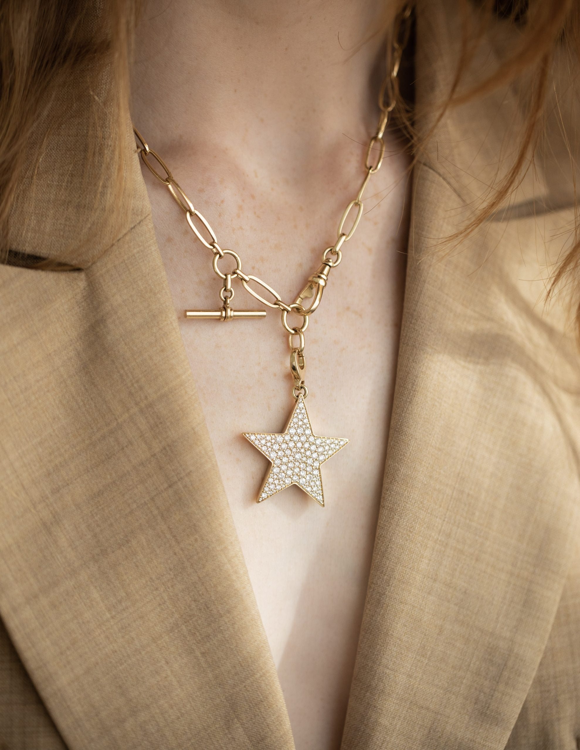 A person in a beige blazer is seen wearing the Jumbo Diamond Star Charm necklace from Janet Heller Fine Jewelry, with custom features like a large star-shaped charm adorned with small gems and a toggle clasp, creating a striking contrast against the blazer.