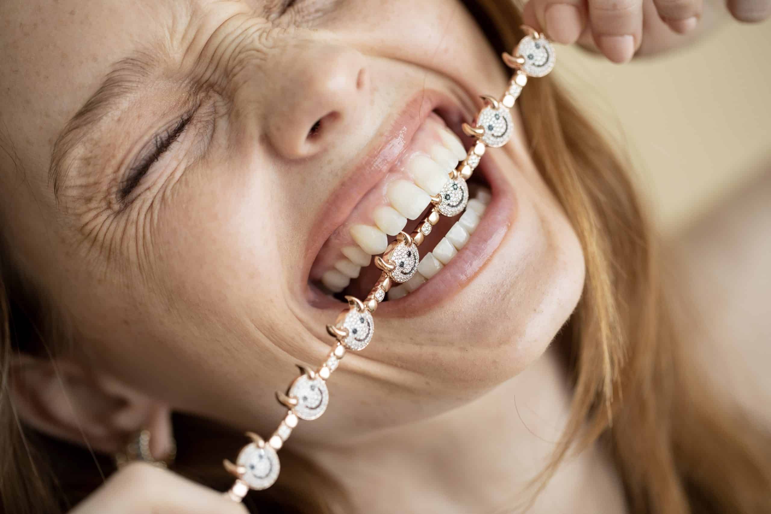 A joyful person with long hair playfully bites a Lucky Devil Bracelet, featuring round decorative elements. Their eyes are closed, savoring its promise of good fortune, while the softly blurred background highlights their face and the striking bracelet.