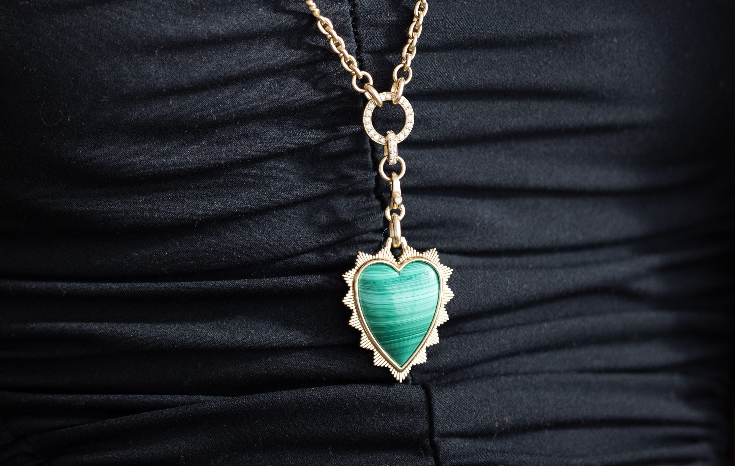 The Malachite Heart Shield Charm by Janet Heller Fine Jewelry showcases a heart-shaped green stone with a striped pattern. It hangs from a gold chain against textured black fabric, featuring an intricate gold border and custom charms for uniqueness.
