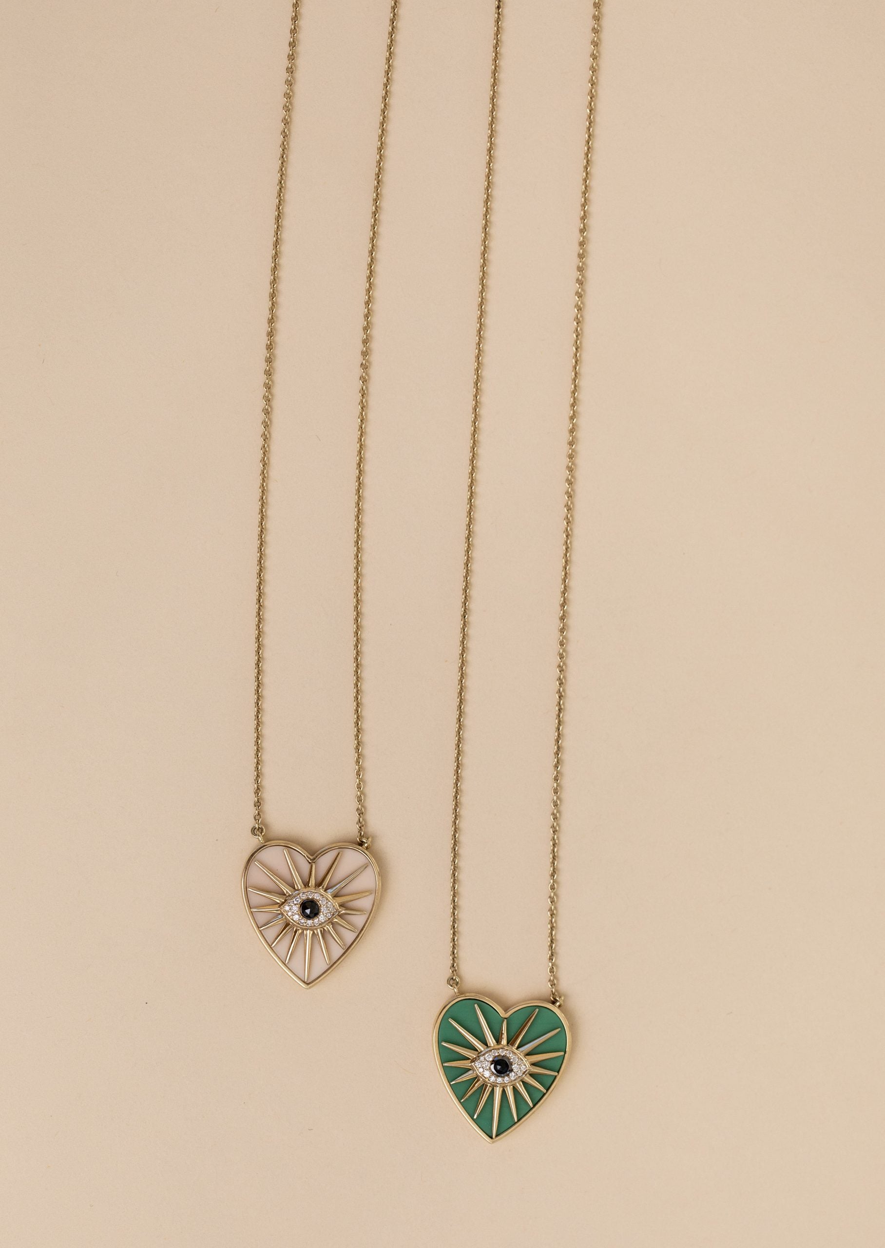 Two Cacholong Heart Burst Necklaces with sunburst designs on gold chains are displayed against a beige background. One pendant is white, the other green, both featuring a central black eye motif. These exquisite pieces are perfect for personal style customizations.